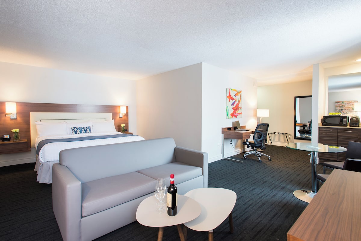 Habitación de hotel con cama, sofá, escritorio con silla y una mesita con botella y vaso.