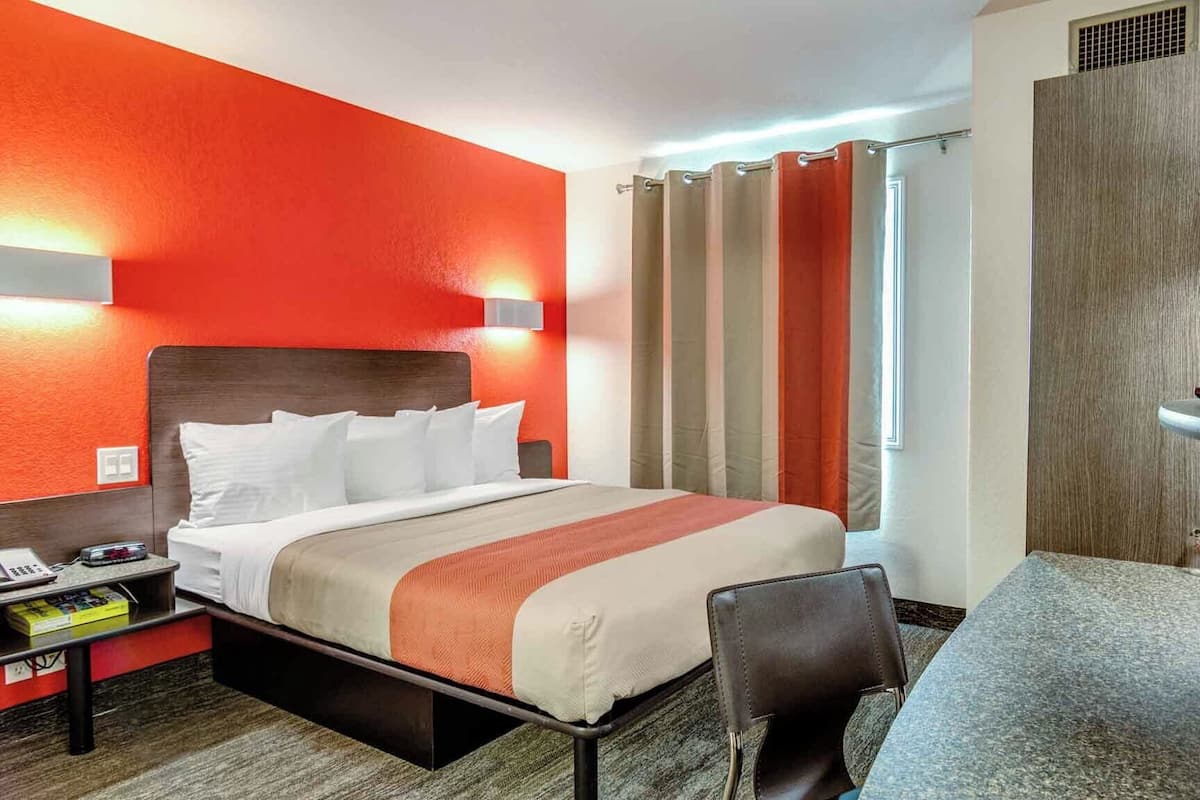 Quarto de hotel com cama, escrivaninha, cadeira, mesa de cabeceira e janela com cortinas.