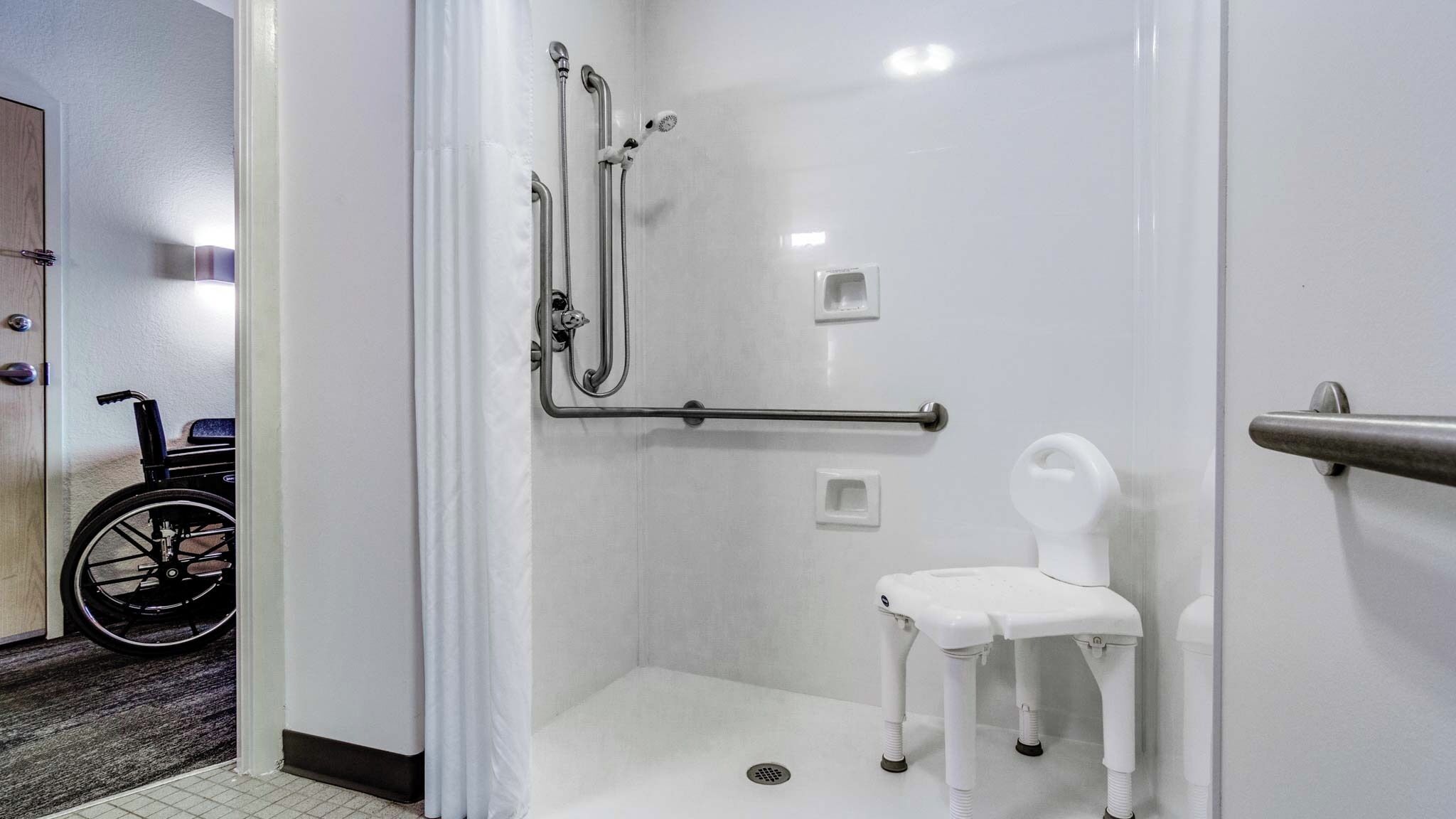 A wheelchair-accessible shower room with a white chair, grab bars, and a handrail.