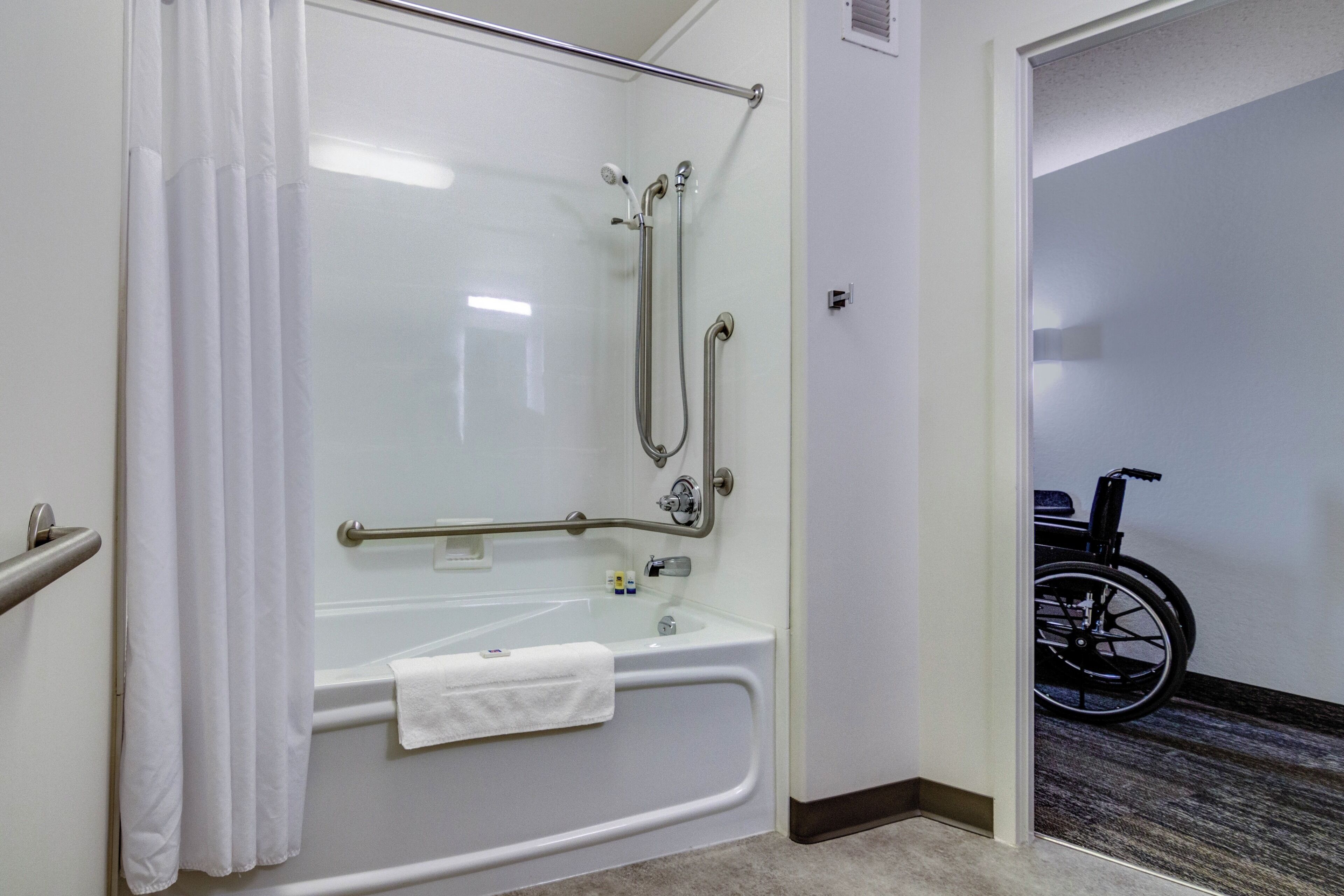 Una doccia accessibile in sedia a rotelle, con barra di sostegno, una vasca da bagno bianca e una sedia a rotelle nella stanza adiacente.