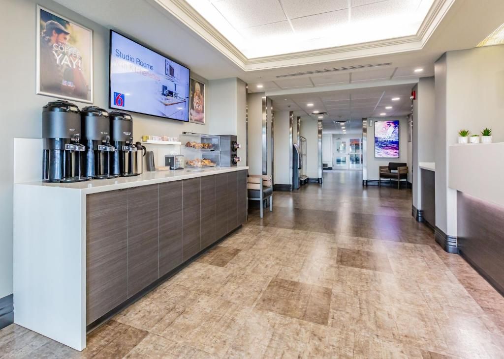 Zona de recepción de un hotel con una estación de café, un televisor mostrando 'Habitaciones Estudio' y un pasillo que conduce a otras áreas del hotel.