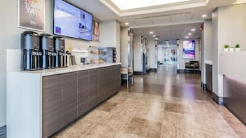 A hotel reception area with a coffee station, a TV displaying 'Studio Rooms,' and a corridor leading to other hotel areas.