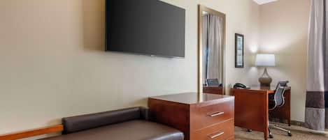 Habitación de hotel con una televisión de pantalla plana montada en la pared, un escritorio de madera con cajones, un banco de cuero, una lámpara de escritorio y un teléfono.