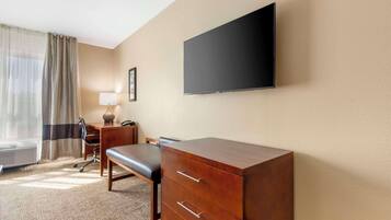 A hotel room with a large flat-screen TV mounted on the wall, a wooden desk with drawers, a cushioned bench, a lamp, and a framed picture.