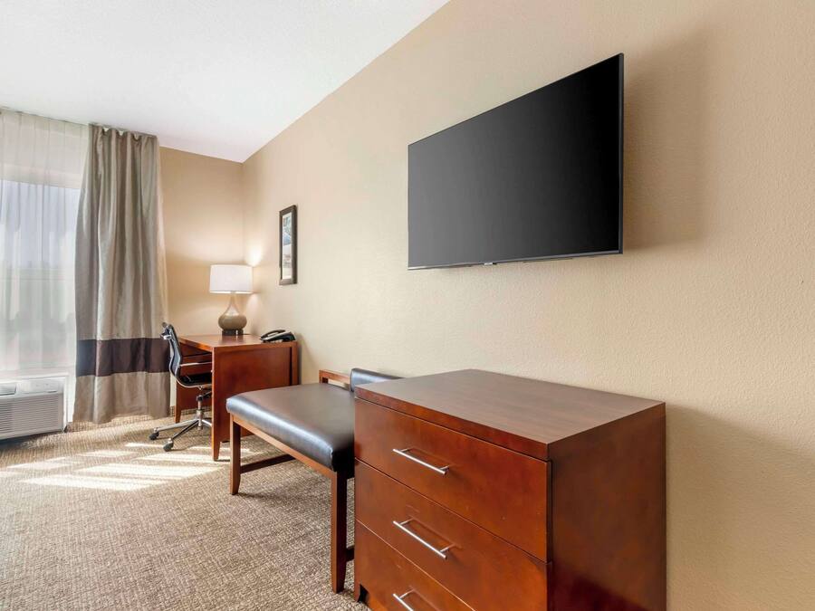 A hotel room with a large flat-screen TV mounted on the wall, a wooden desk with drawers, a cushioned bench, a lamp, and a framed picture.