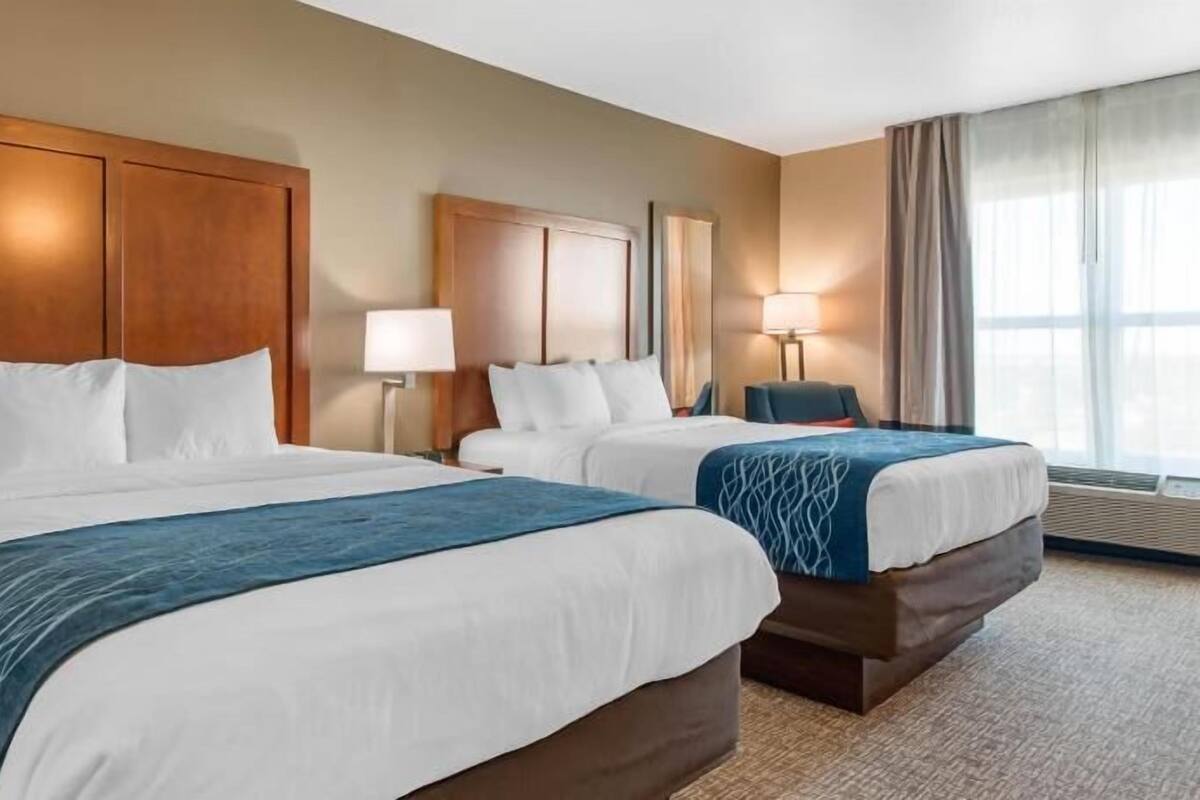 A hotel room with two beds, a desk, a chair, a lamp, and a large window.