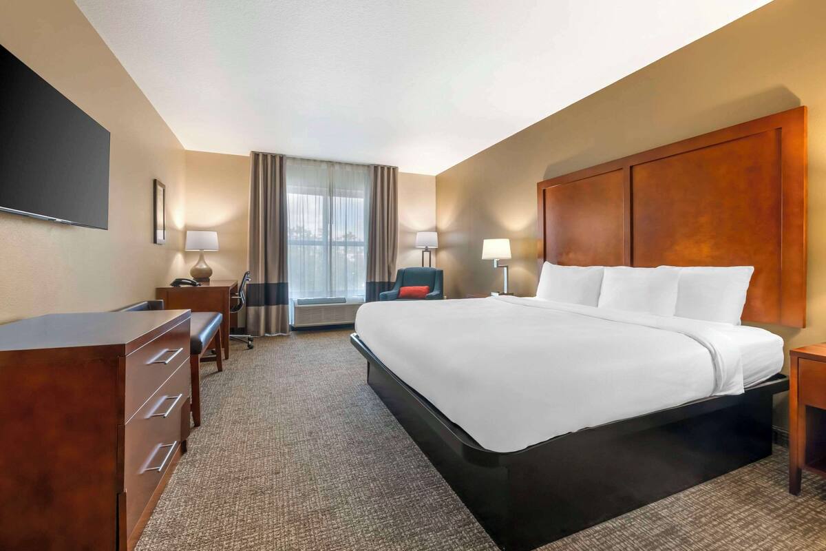 A hotel room with a large bed, a desk with drawers, a chair, a TV mounted on the wall, and a window with curtains.
