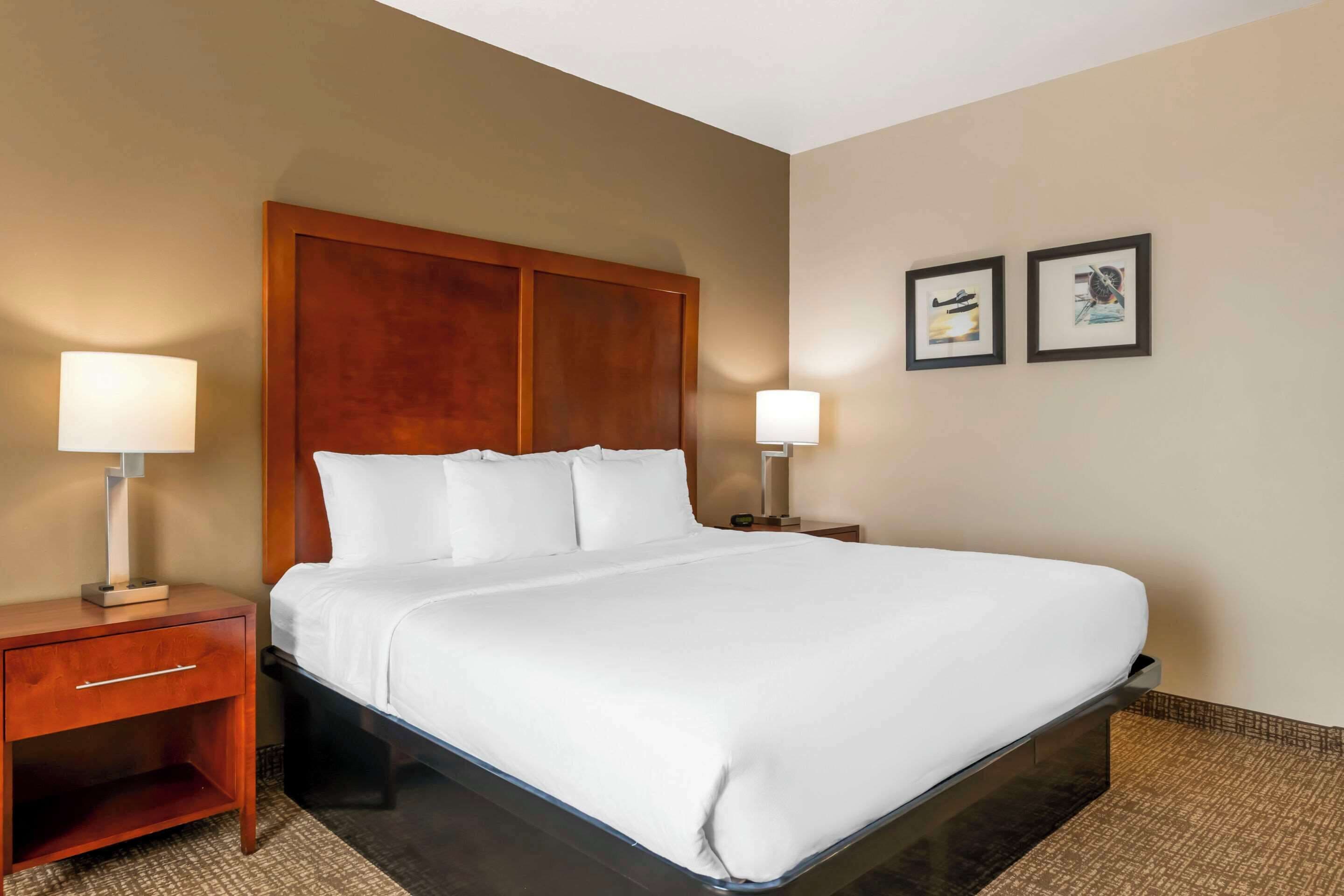 Una habitación de hotel con una cama grande, cabecero de madera, dos mesitas de noche con lámparas y dos cuadros enmarcados en la pared.