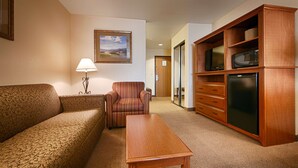 Habitación de hotel con un sofá estampado, un sillón a rayas, una mesa de centro de madera, un televisor sobre un mueble de madera y un cuadro con paisaje en la pared.
