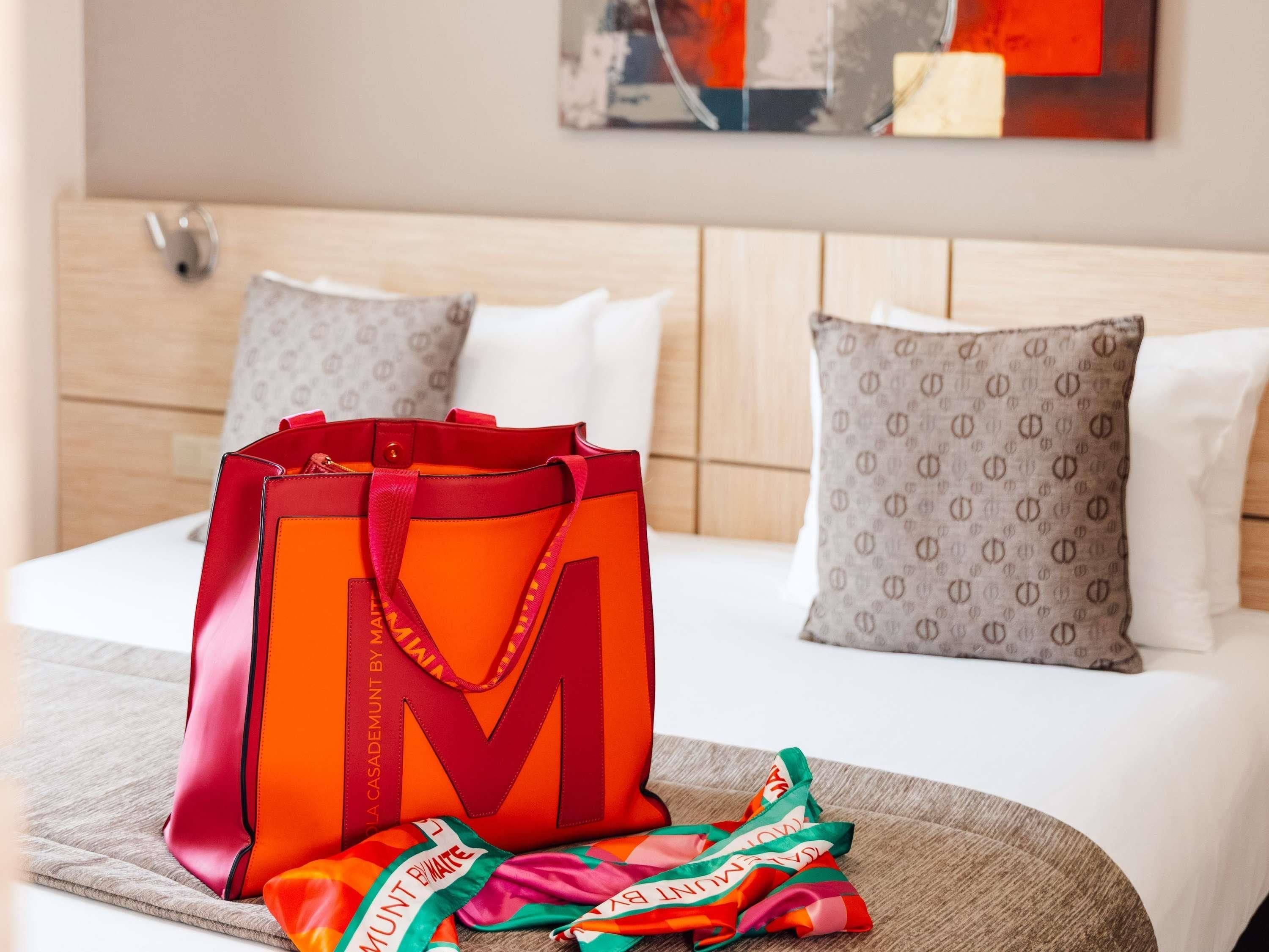 Een hotelkamer met een bed, kussens, een deken, een grote oranje tas en een kleurrijke sjaal.