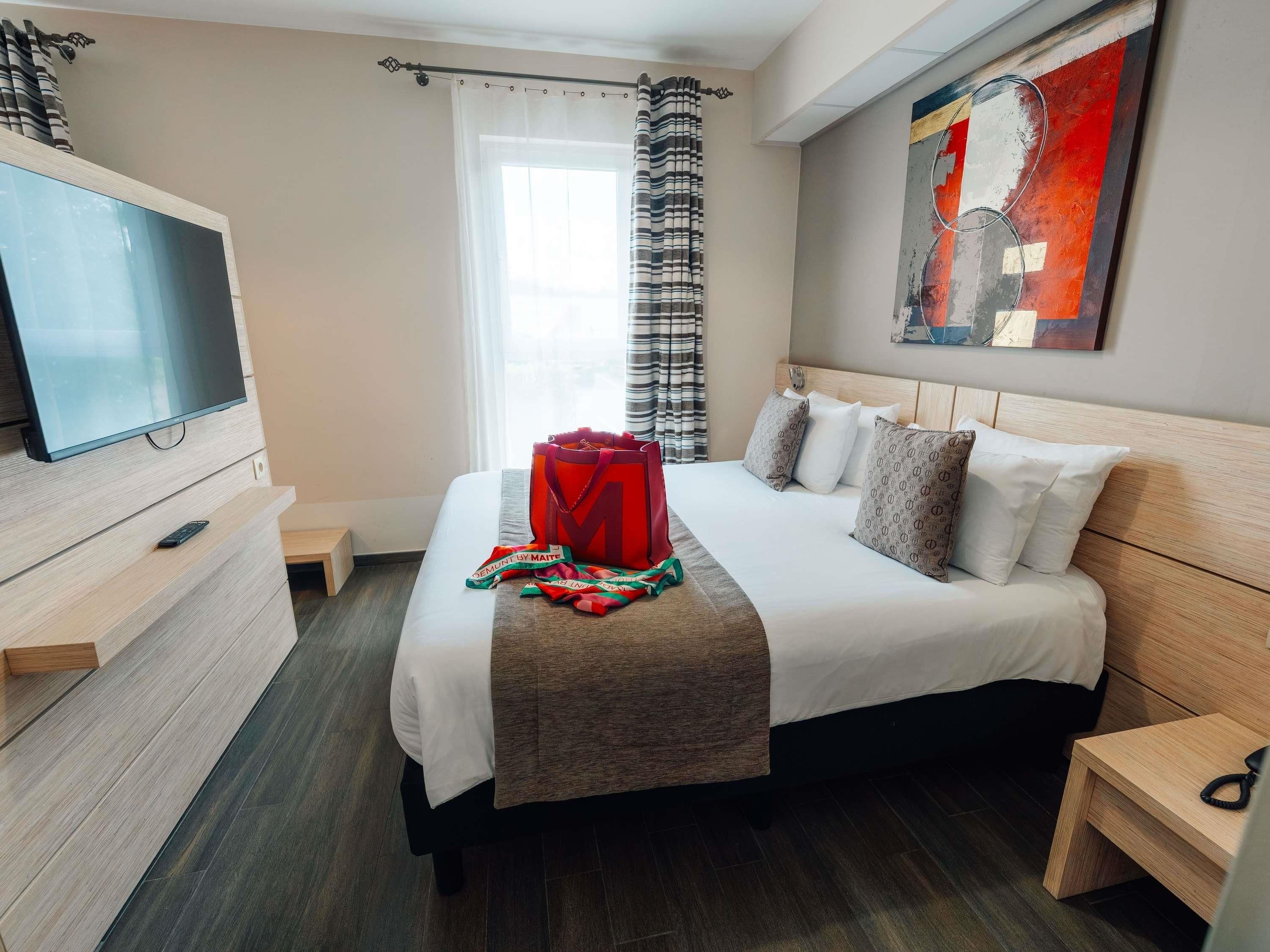 A hotel room with a bed, a TV, a painting, and a window with curtains.