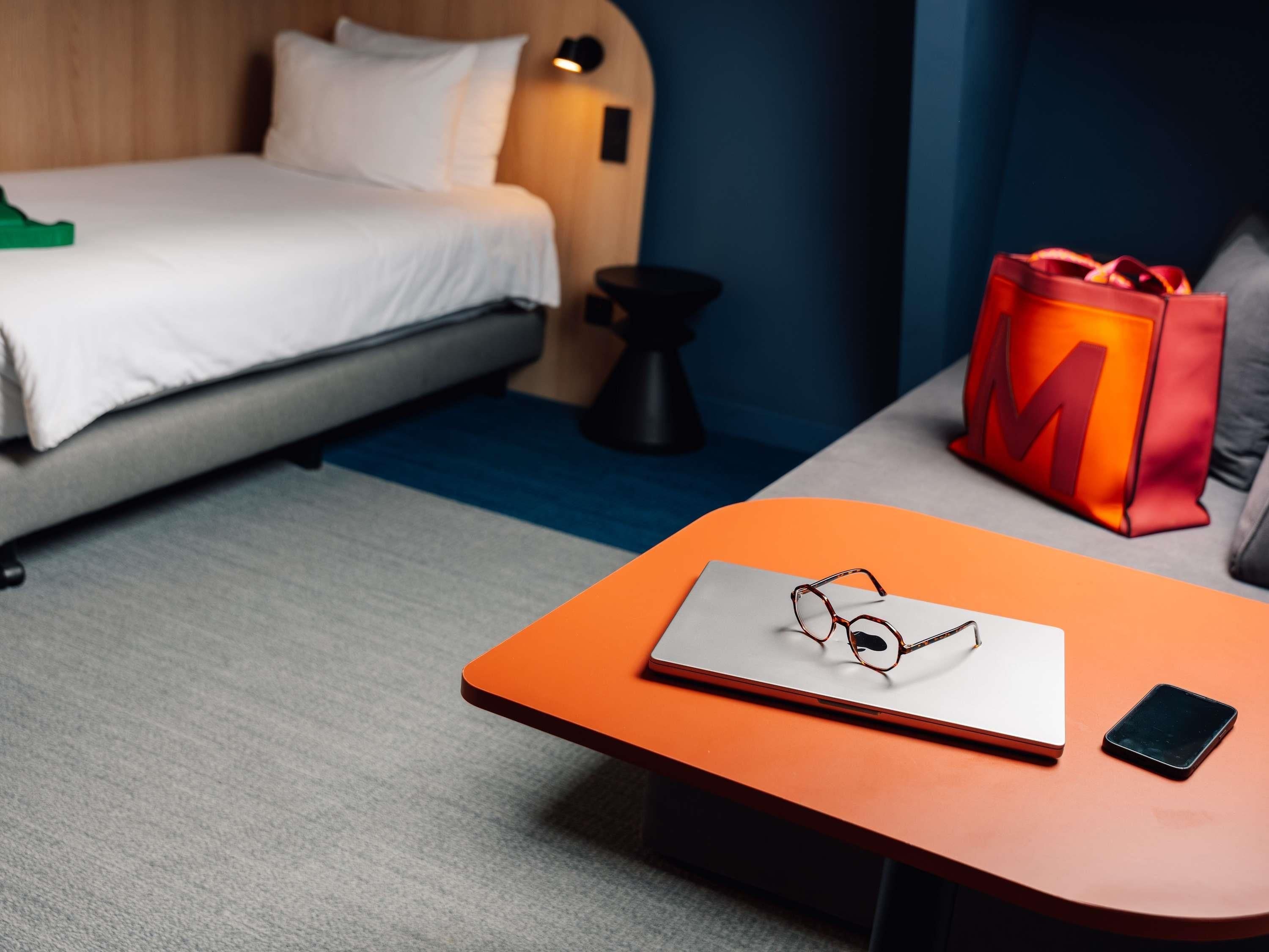 A hotel room with a bed, a small table with a laptop, glasses, and a smartphone.