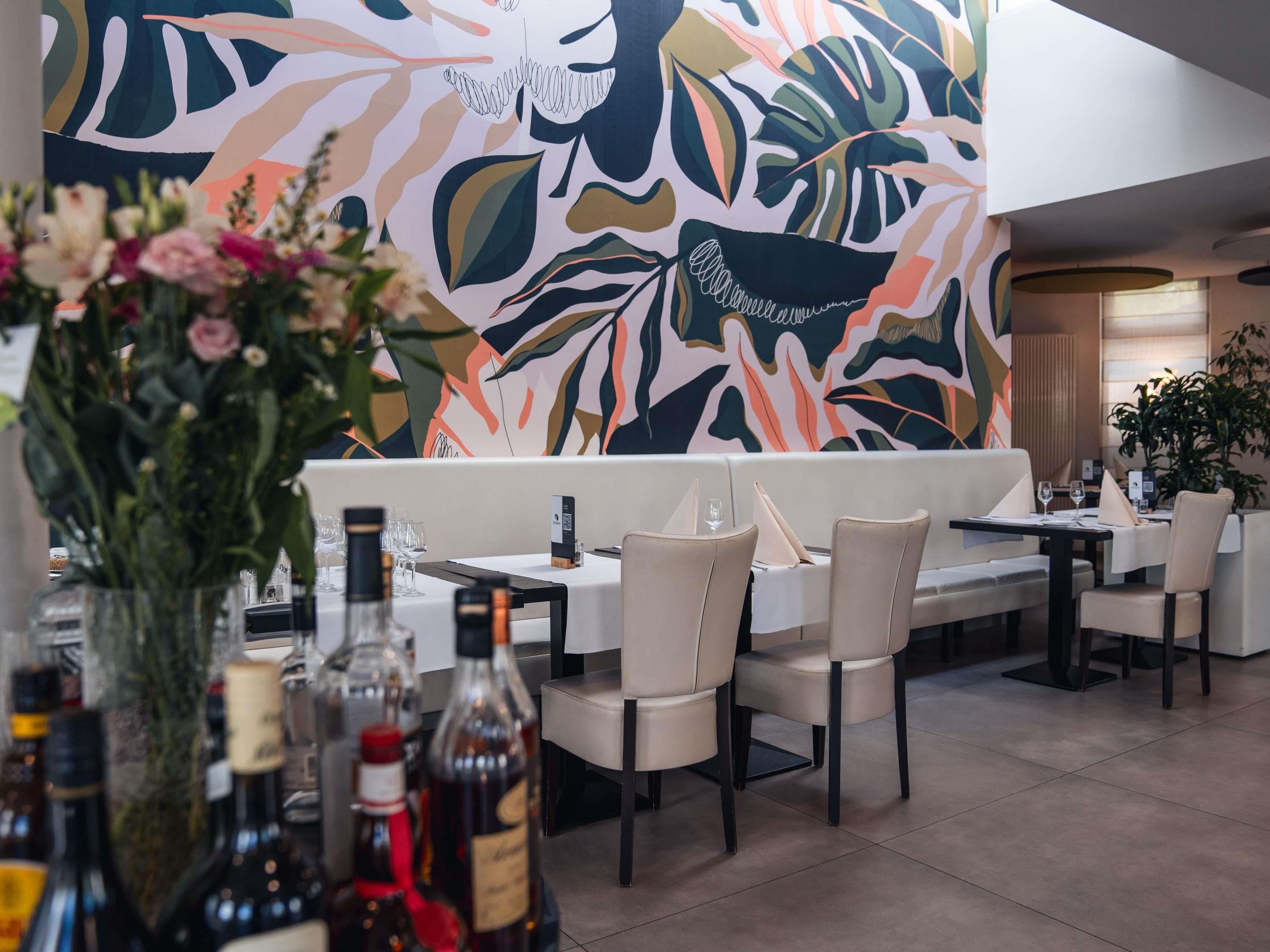 A restaurant interior with a modern design, featuring a large abstract mural on the wall, a seating area with tables and chairs, and a vase with flowers on the table.