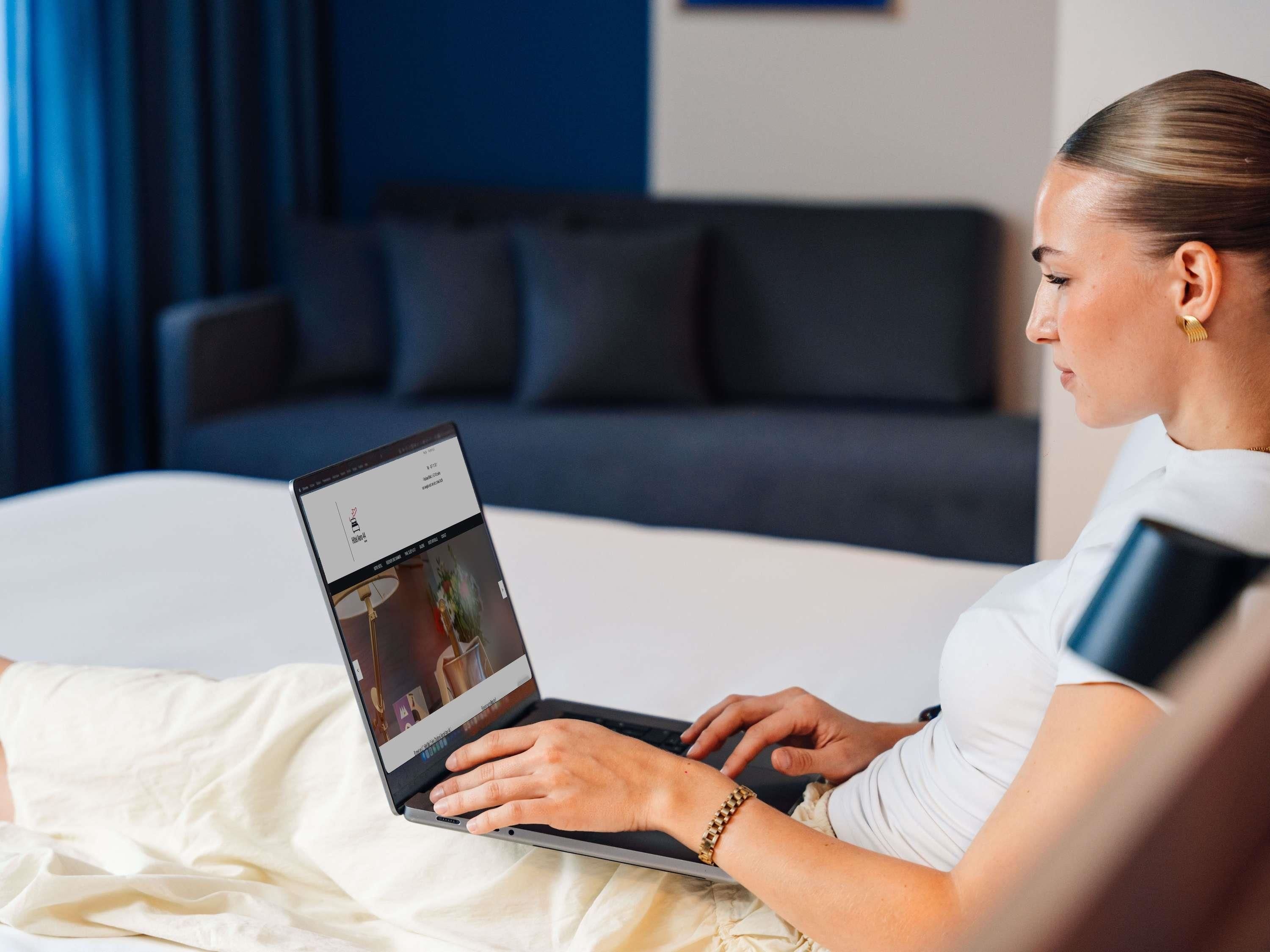 Una persona usando una computadora portátil en una cama, en una habitación con un sofá y cortinas oscuras.
