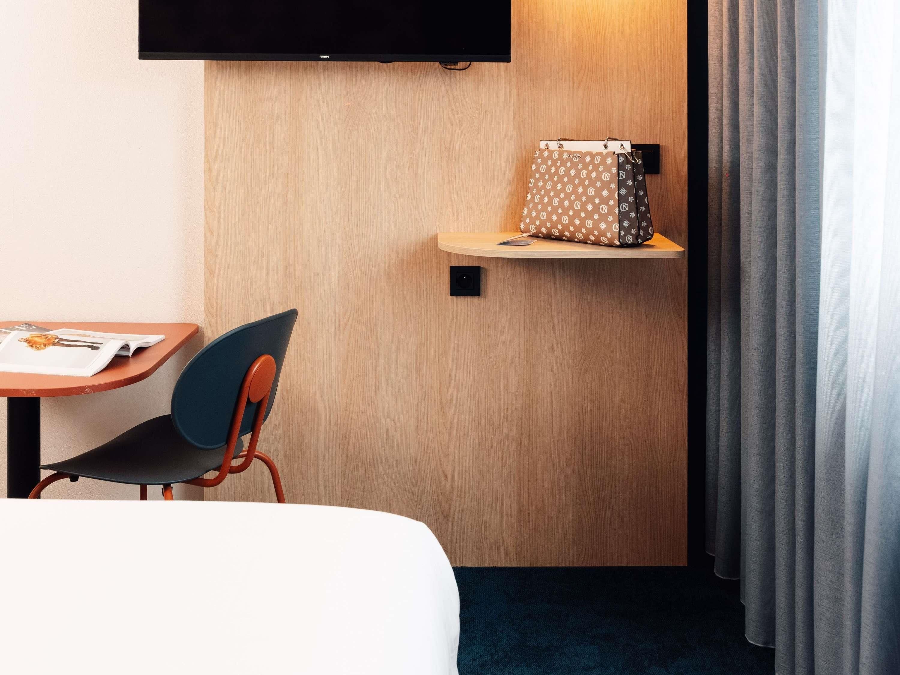 A hotel room with a bed, a desk with a chair, a television mounted on the wall, a small shelf with a bag, and a window with curtains.