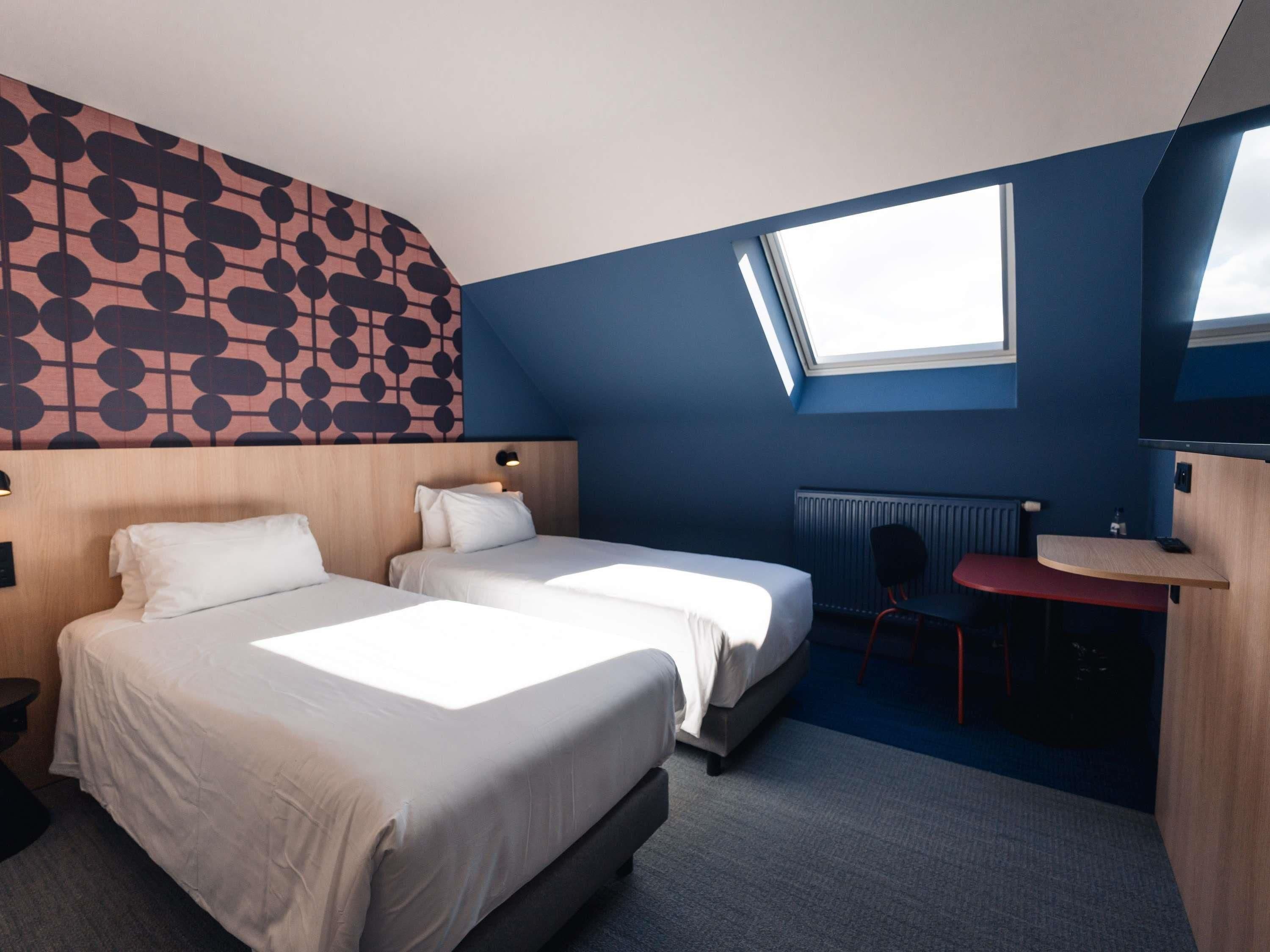 Habitación de hotel con dos camas, un escritorio, una silla, un radiador y un tragaluz.