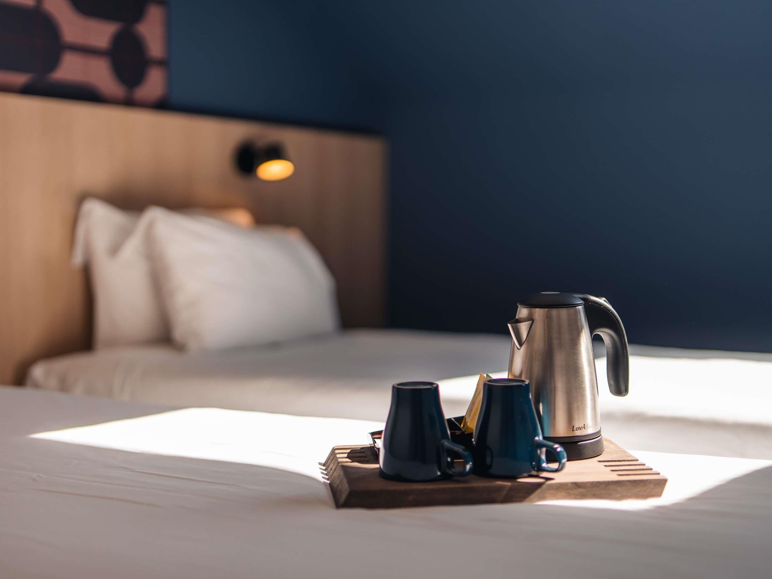 A neatly made bed with a tray of coffee mugs and a kettle on a wooden board.