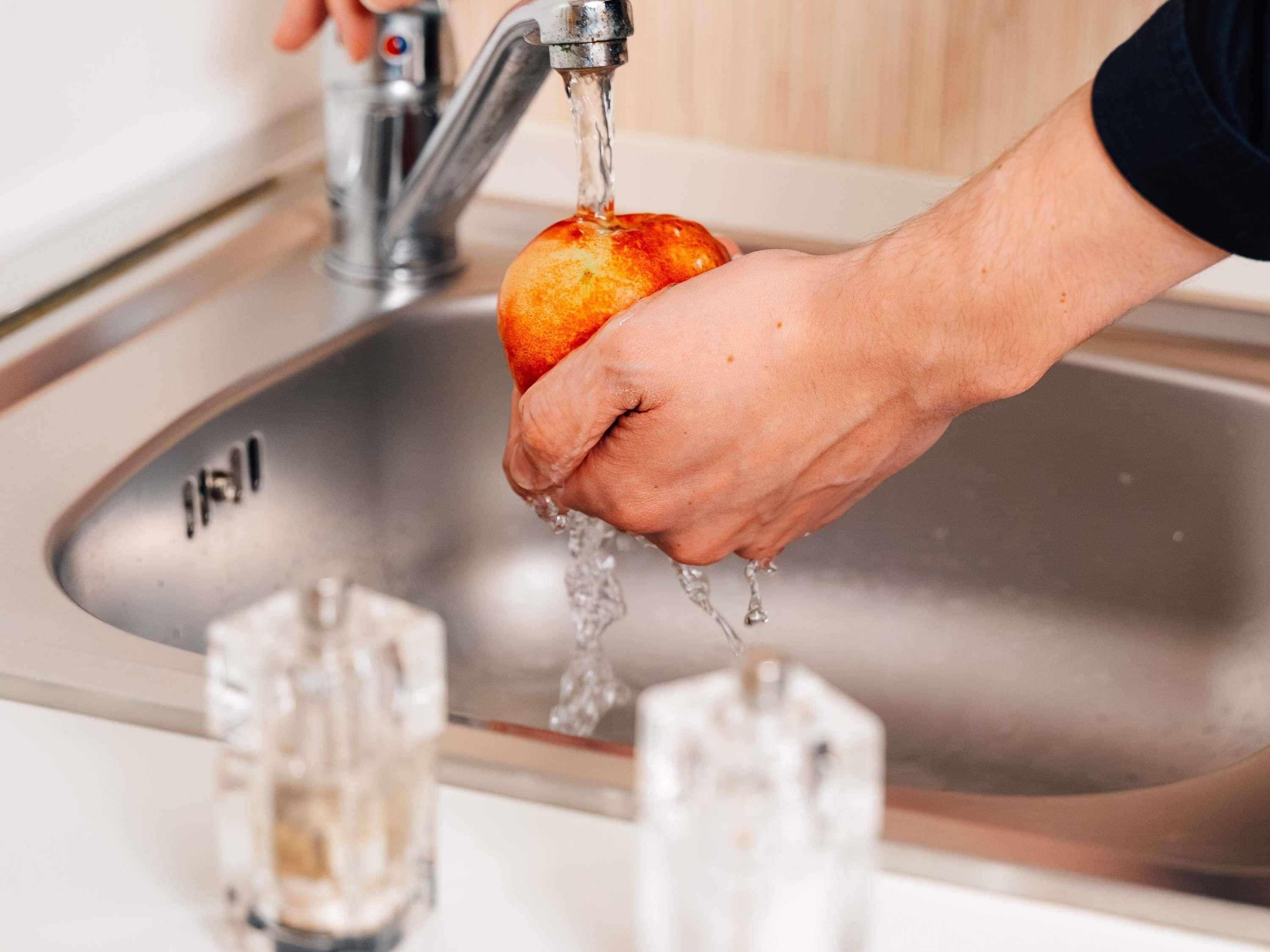 A hand holding an orange under a running faucet in a kitchen sink.