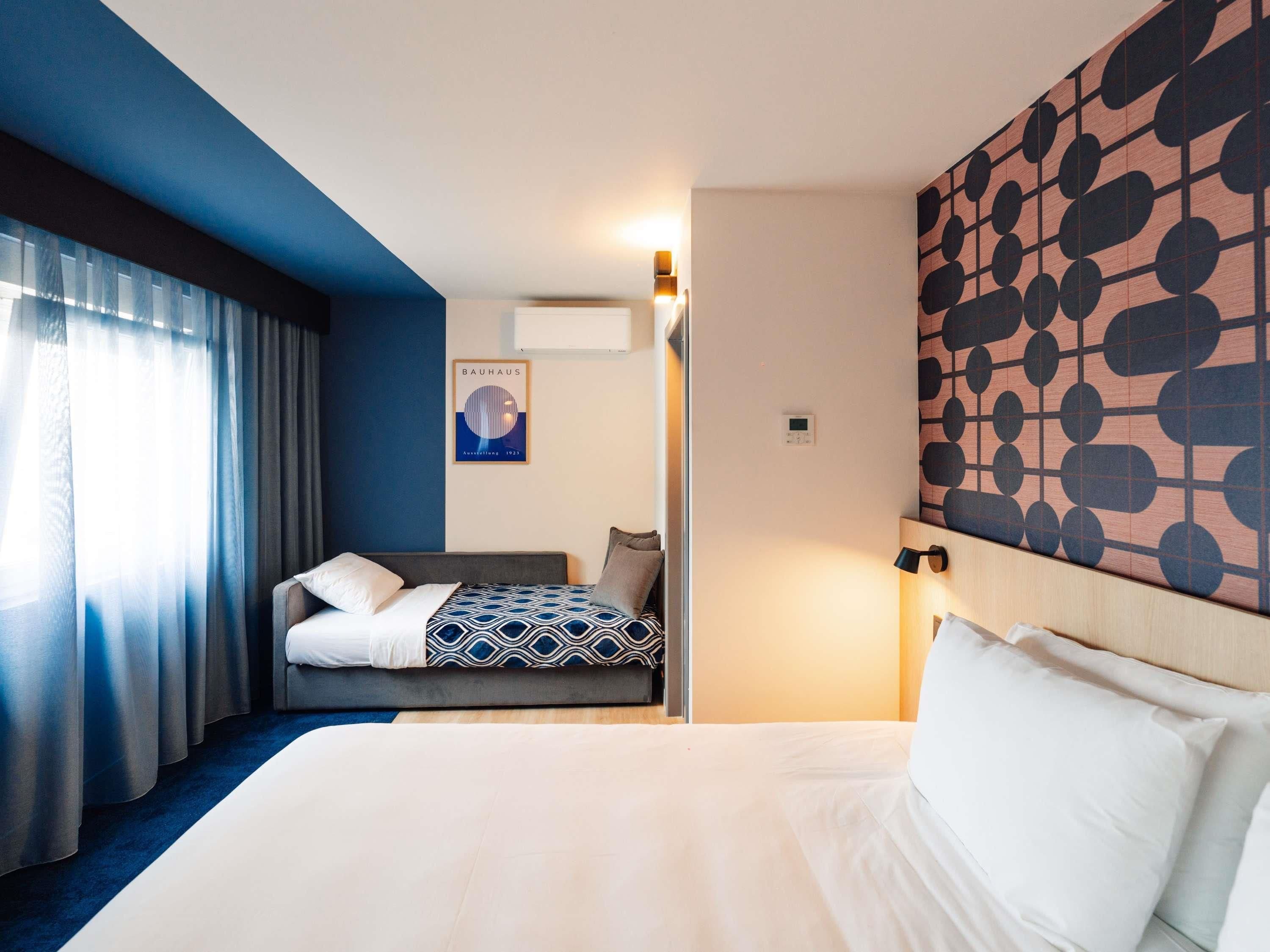 Une chambre d’hôtel avec un grand lit, un plus petit lit, un climatiseur fixé au mur, une affiche et une tête de lit à motifs.