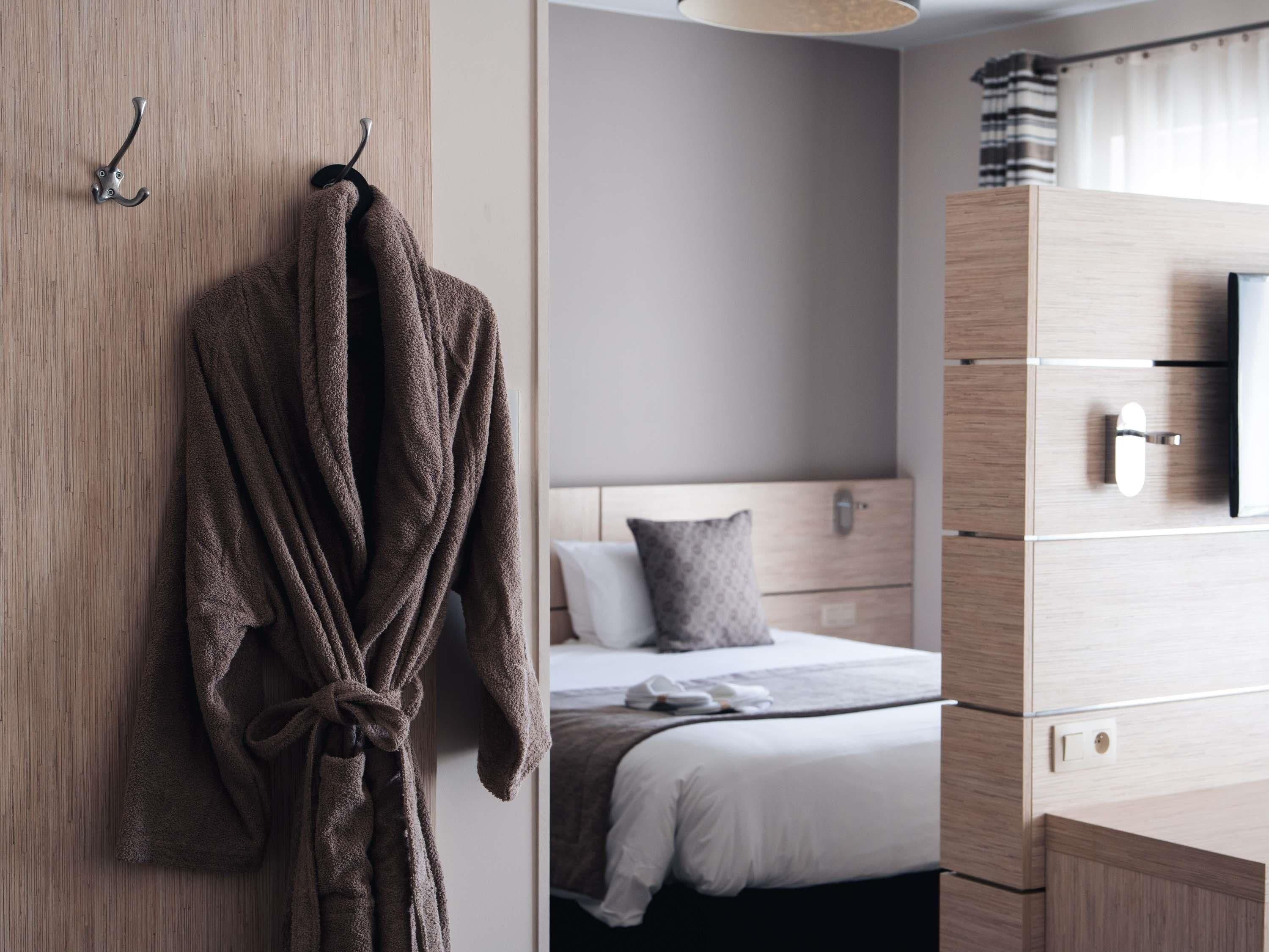 Una habitación de hotel con un armario de madera, una cama con edredón blanco, una almohada gris y una bata marrón colgada de un perchero.