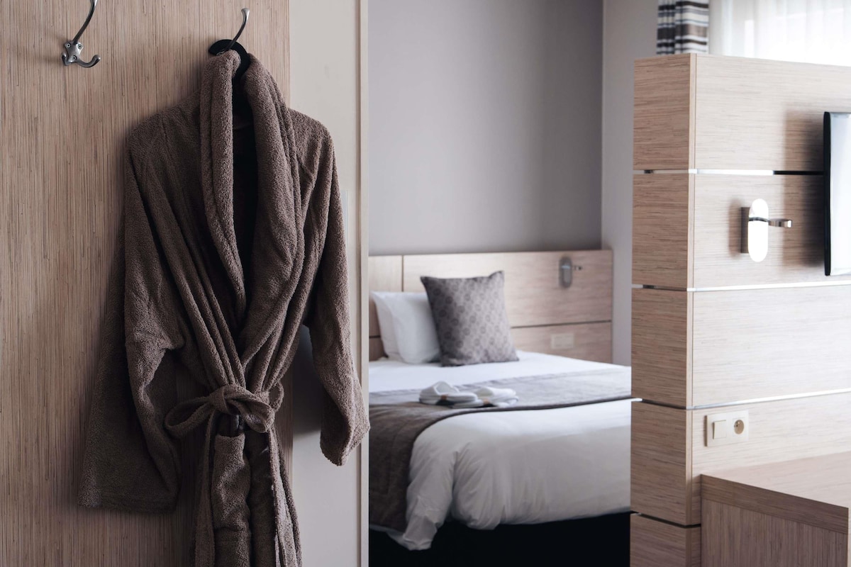A hotel room with a wooden wardrobe, a bed with a white duvet, a grey pillow, and a brown robe hanging on a hook.