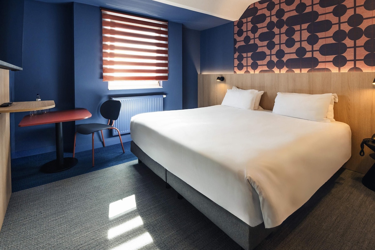 A hotel room with a large bed, a desk, a chair, a radiator, and a patterned wall.