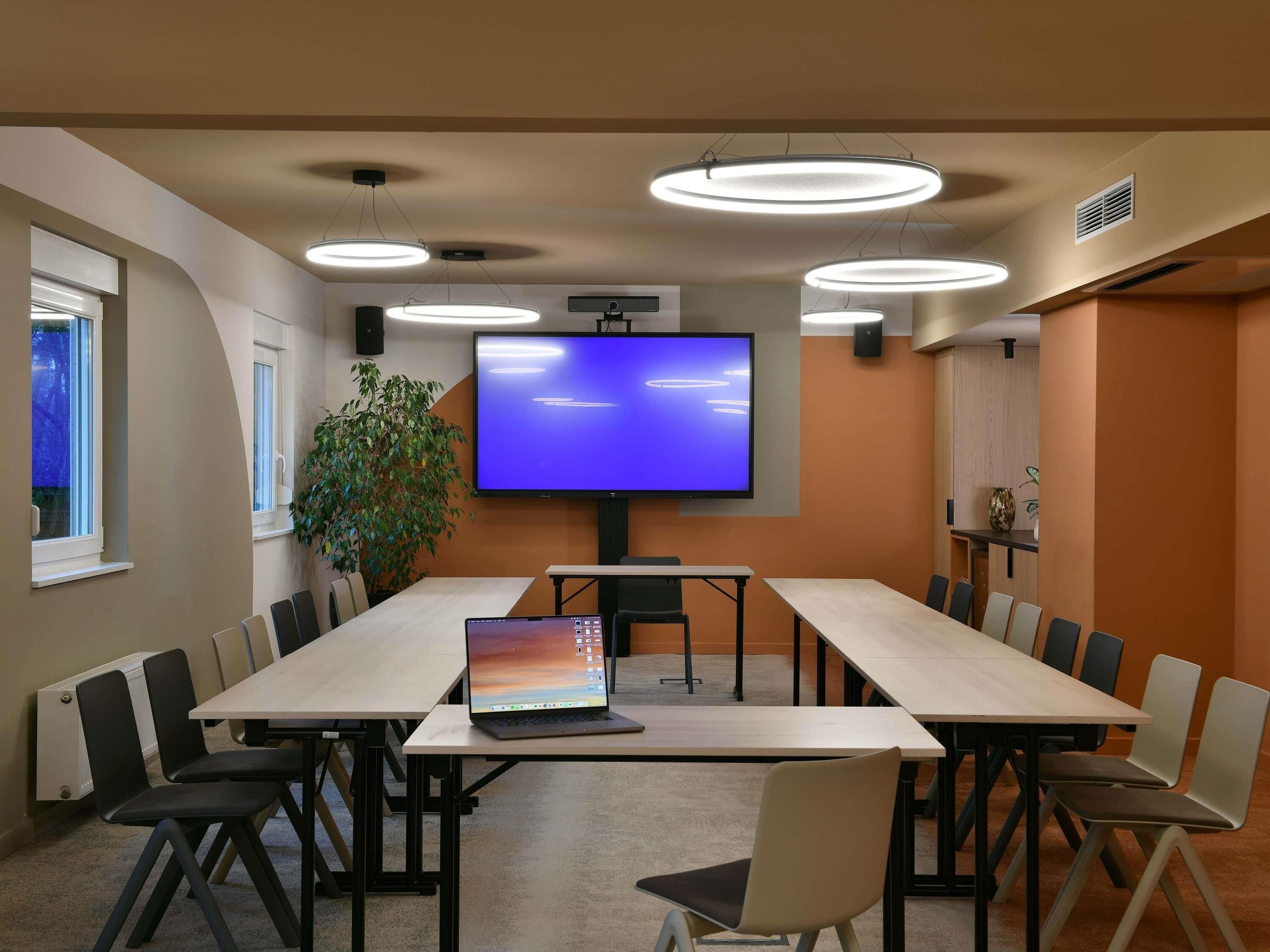 A conference room with a long table, chairs, a large screen, and ceiling lights.