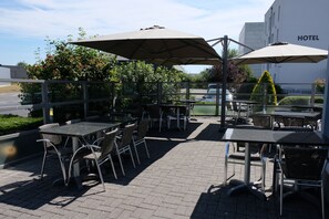 An outdoor patio with tables and chairs, umbrellas, and a view of a hotel building in the background. - Ibis Styles Charleroi Airport Aero44 (Charleroi)