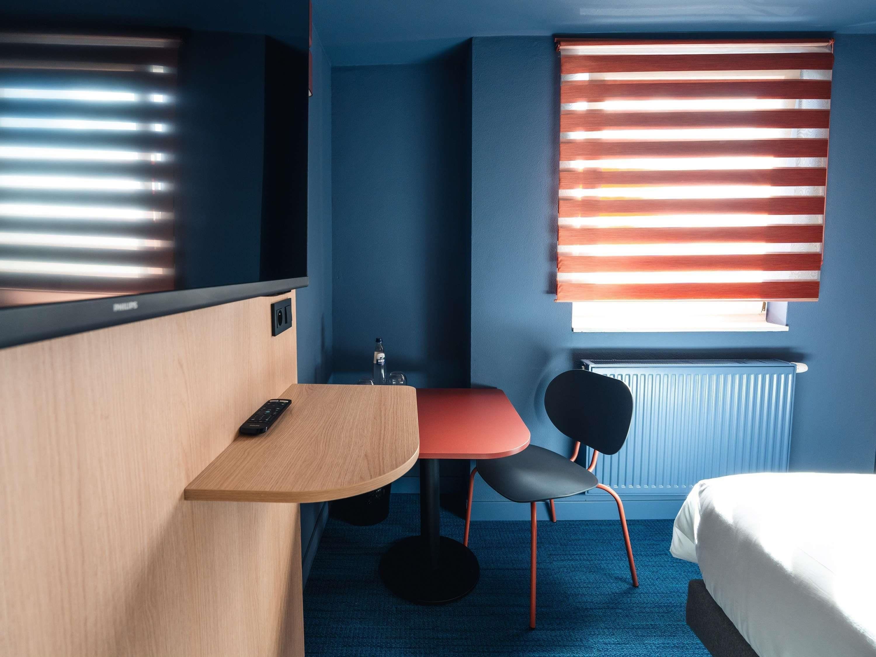 A hotel room with a bed, a desk with a remote control, a chair, a window with blinds, and a radiator.