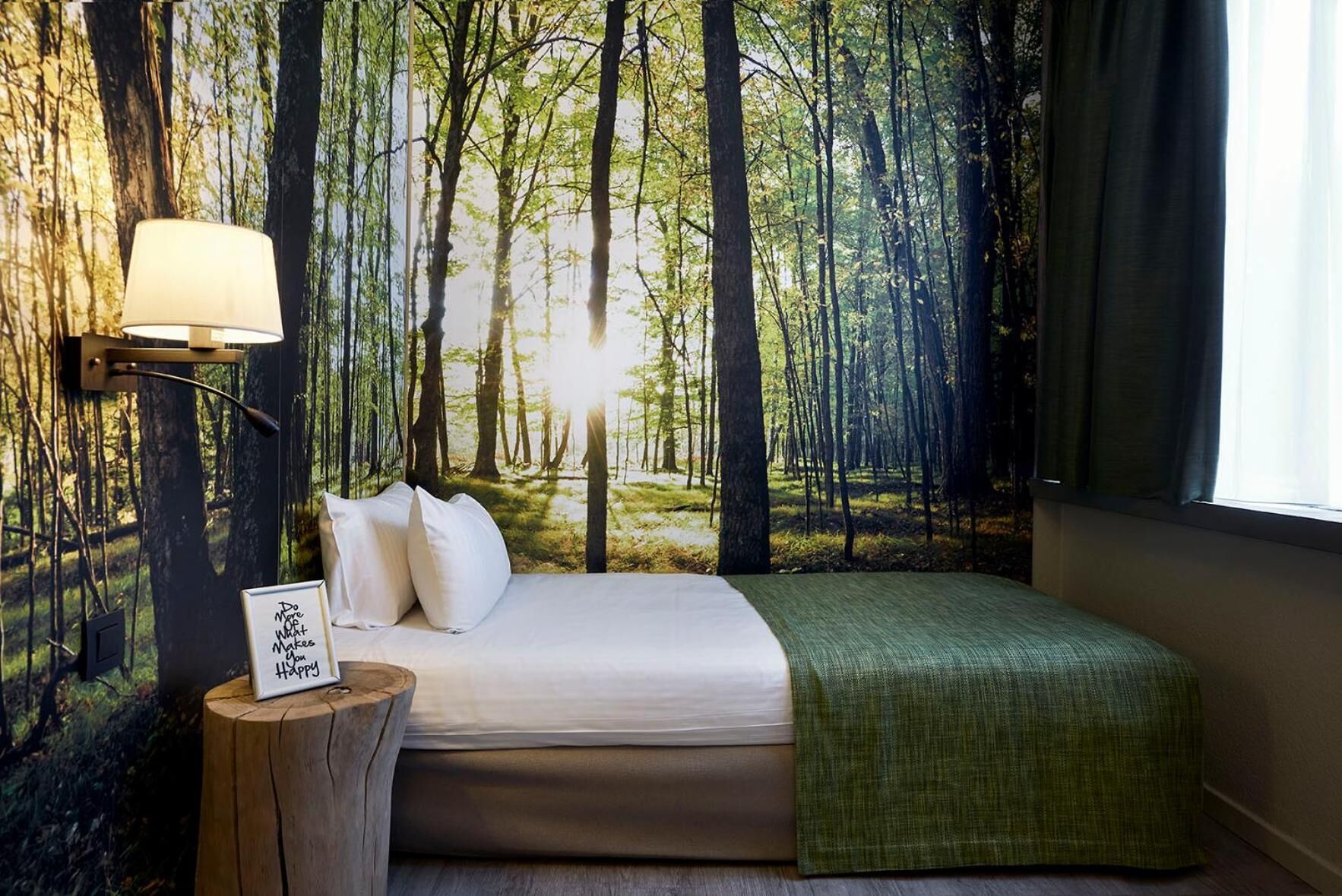 Un dormitorio con un mural temático de bosque, una cama con sábanas blancas, un plaid verde y una lámpara de noche.