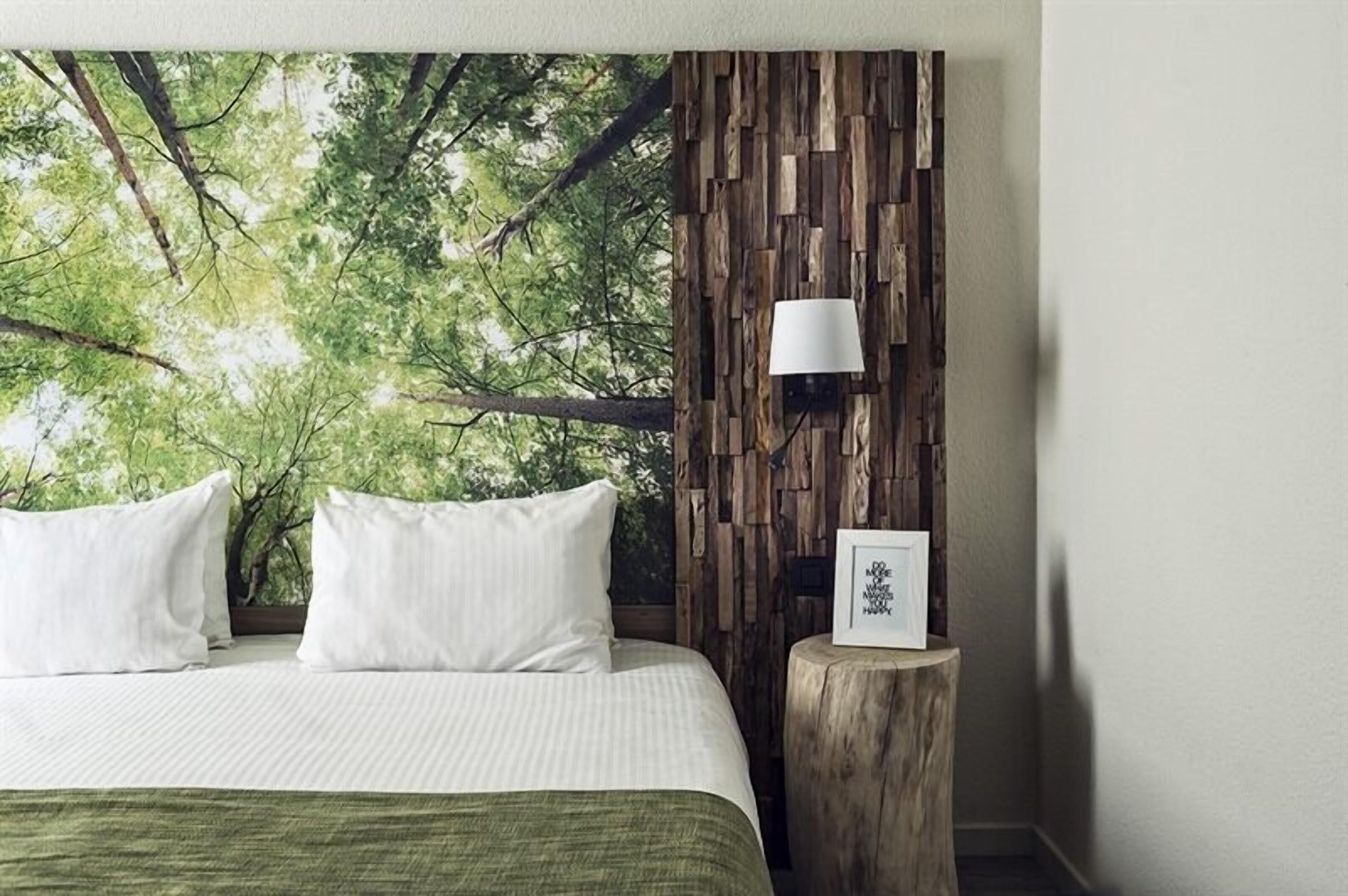 A bedroom with a bed, white pillows, a wooden headboard, a wall-mounted lamp, and a framed picture on a wooden stool.