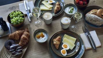Desayuno con cruasanes, huevos, yogur y fruta en platos, junto con vasos de vino y café.