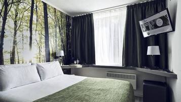 A hotel room with a bed, a window with curtains, a wall-mounted TV, and a bedside table with a lamp.
