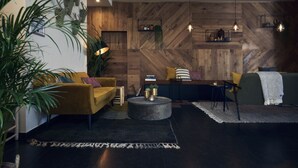 Un moderno salón con una pared de madera en patrón de espiga, un sofá amarillo, un sofá verde, una mesa de centro redonda e iluminación colgante.