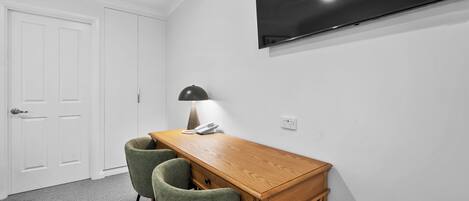 Una habitación con un escritorio de madera, dos sillas verdes, una lámpara, un teléfono y un televisor de pantalla plana montado en la pared.