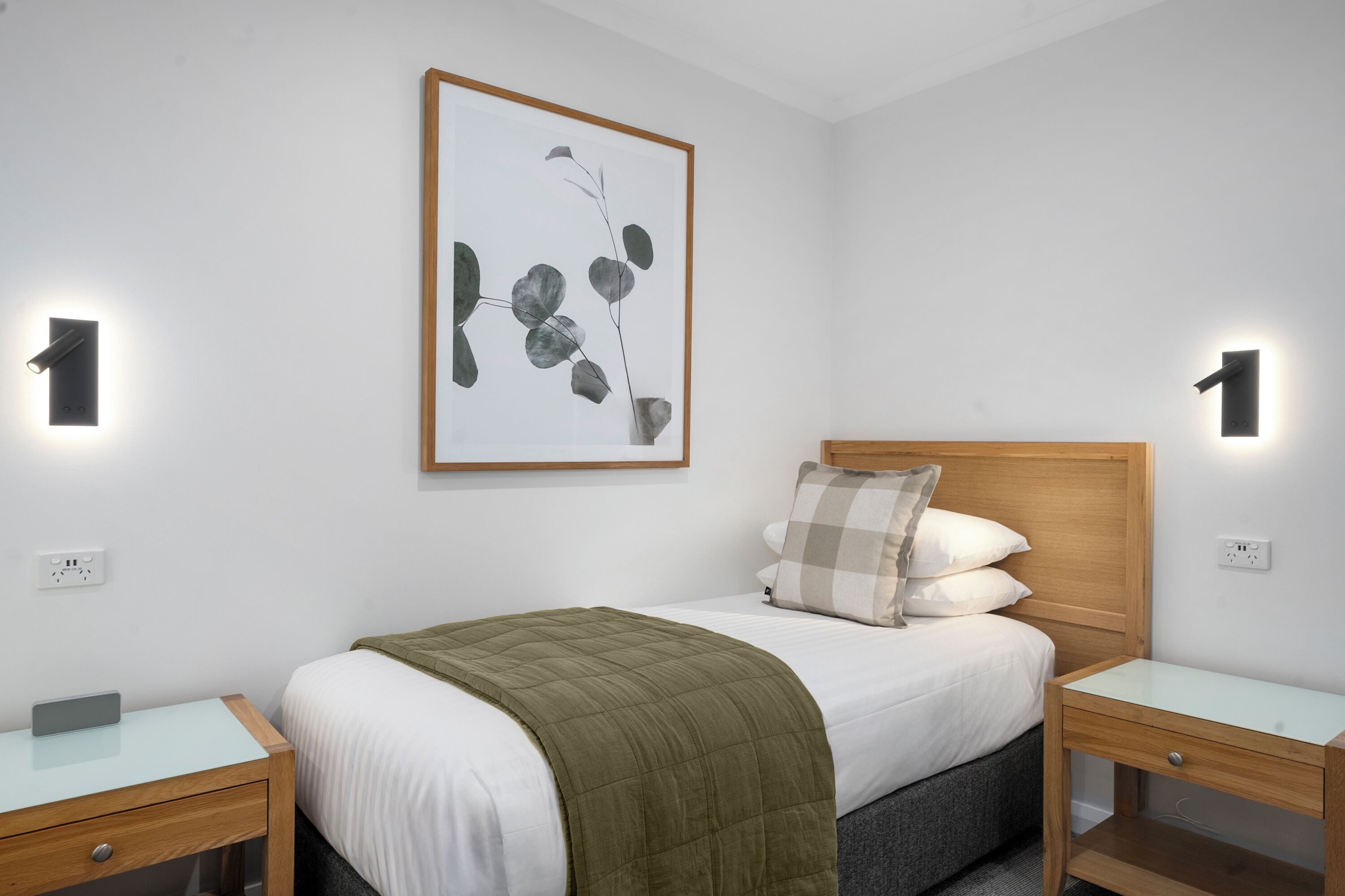 A neatly arranged bedroom with a bed, two bedside tables, a wall-mounted light, and a framed artwork.
