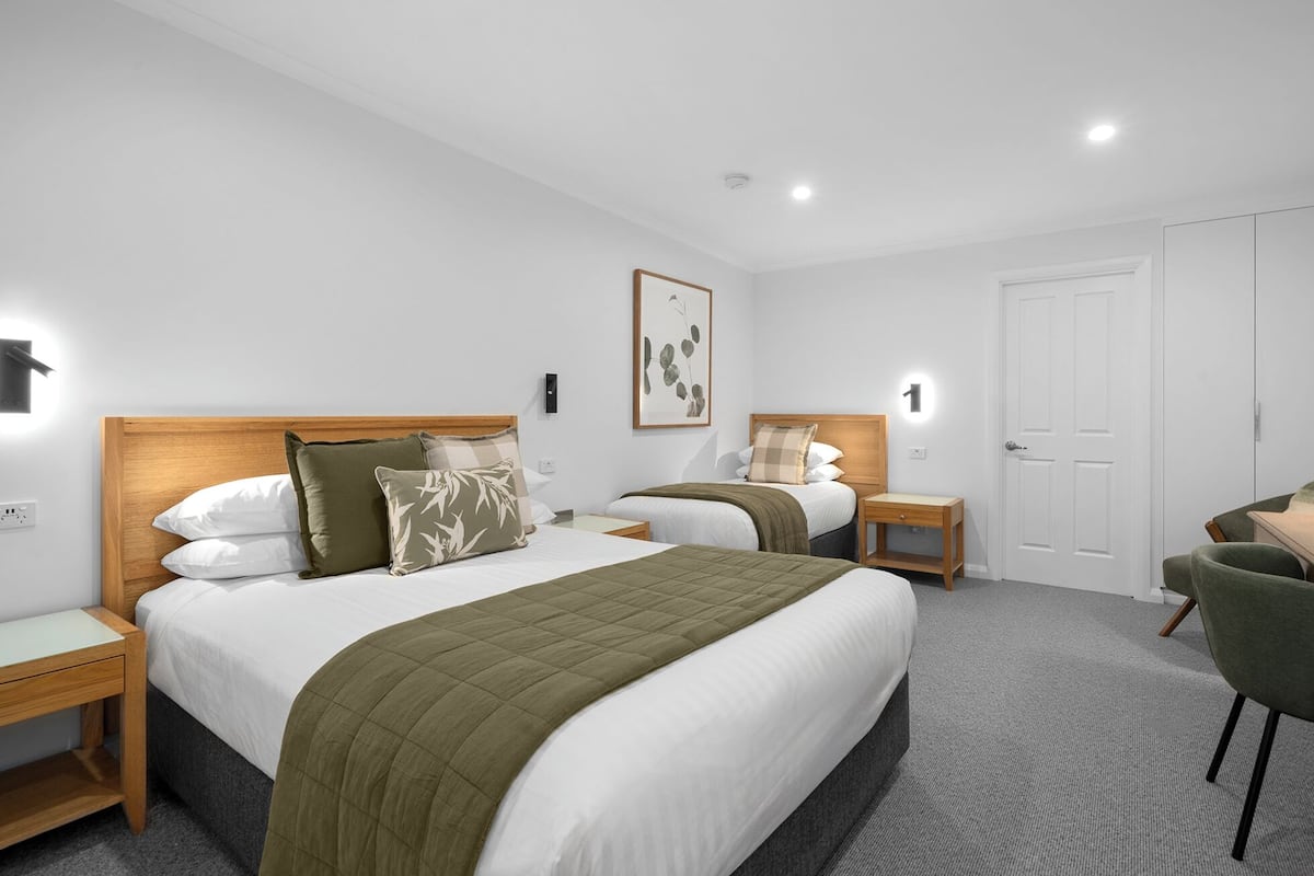 A hotel room with two beds, a desk, and a chair. There are bedside tables with lamps, a wall-mounted light, and a door.