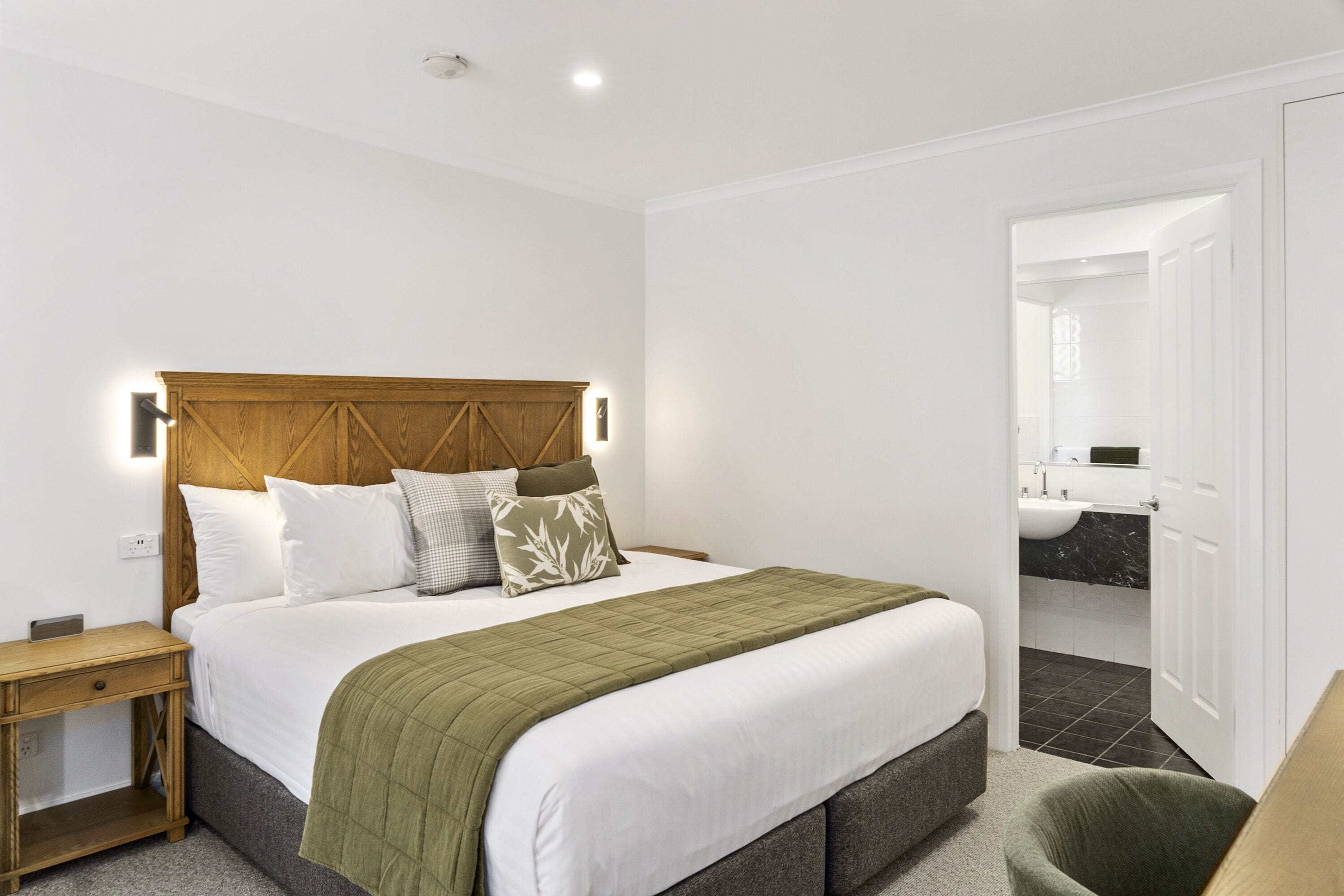 A bedroom with a wooden headboard, a bed with white bedding and a green throw, a bedside table, and a view into the bathroom.