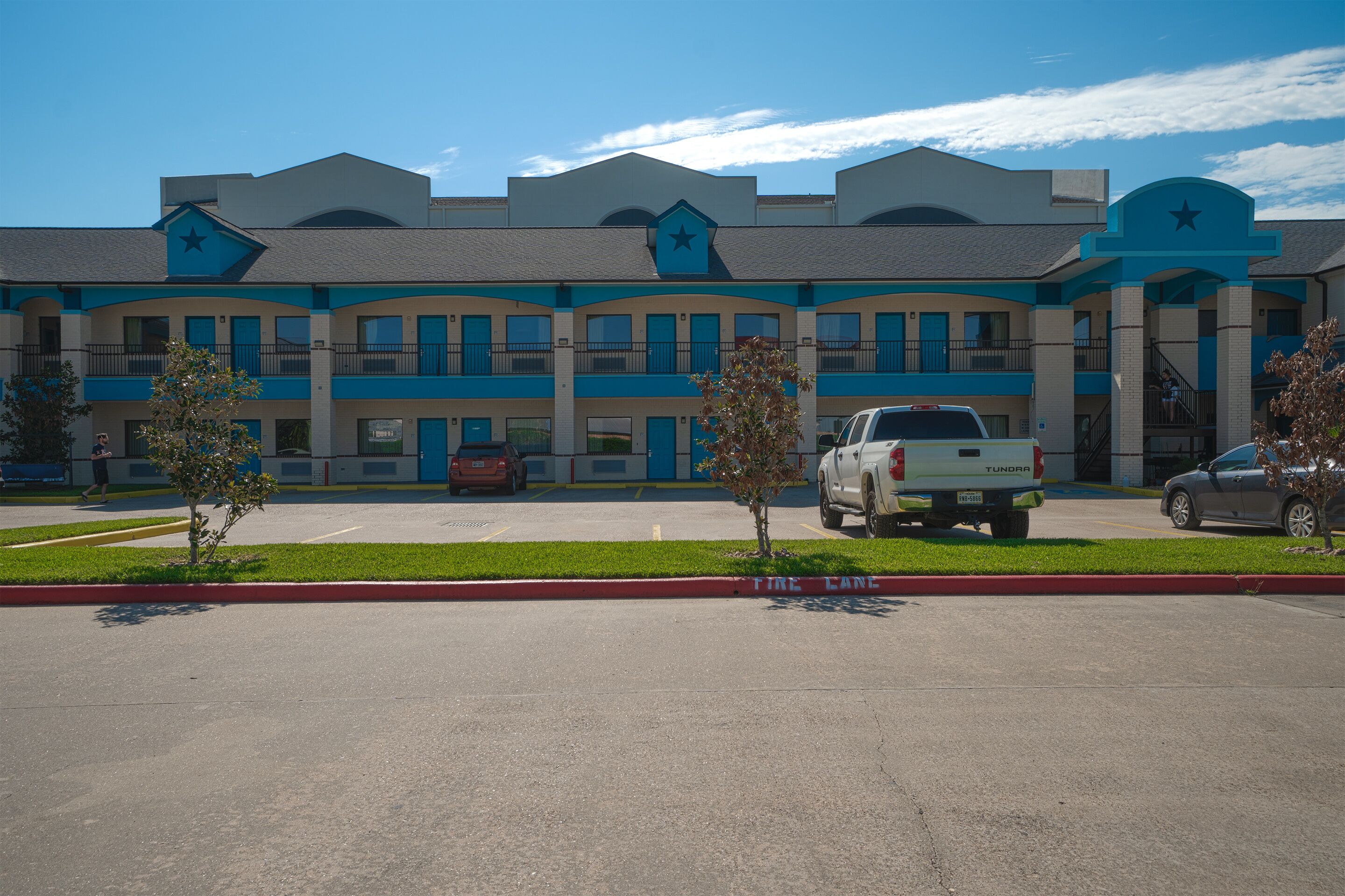 A multi-story hotel with a blue and beige color scheme, featuring a central entrance with a star motif, and a parking lot with several vehicles.
