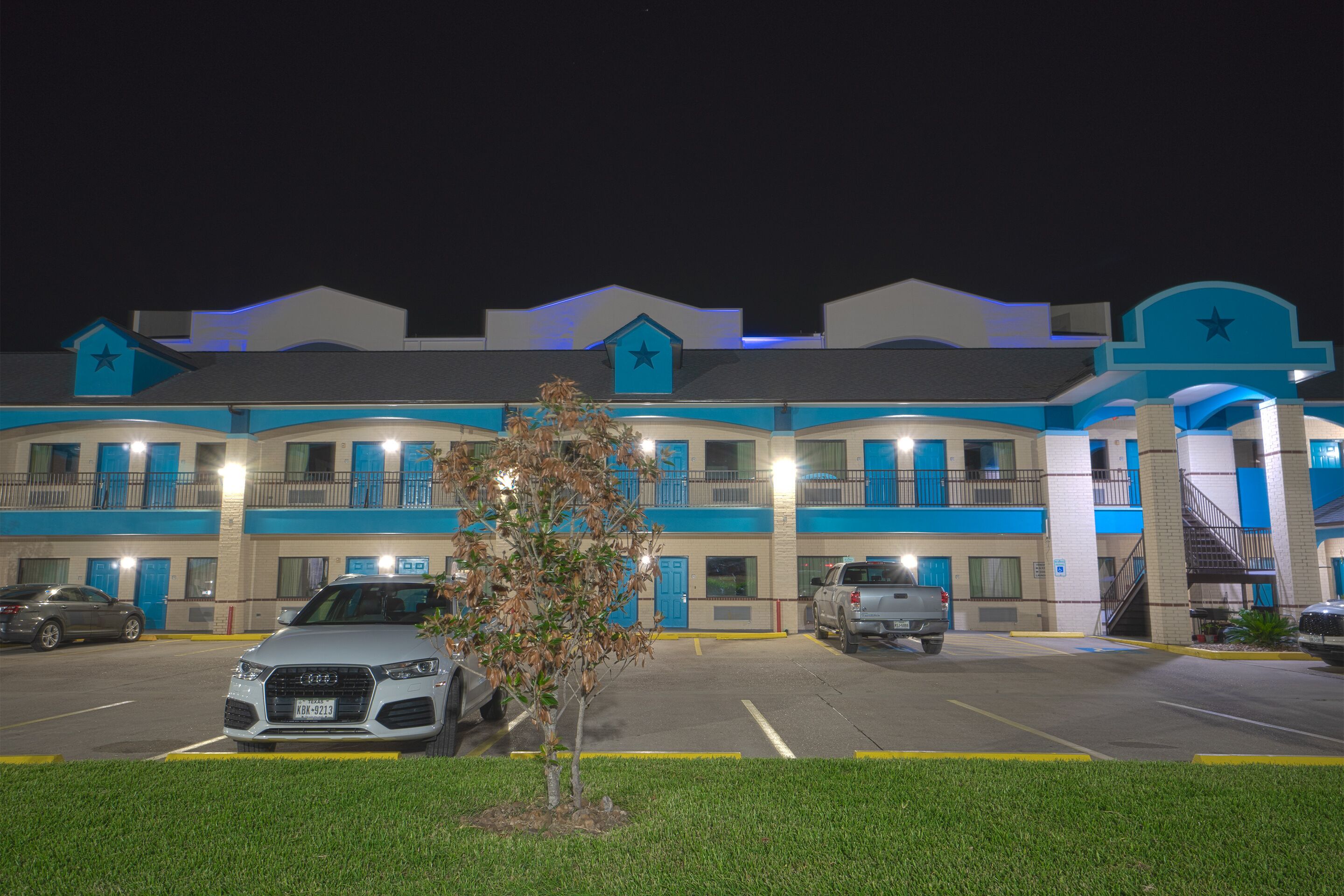 Un hotel con fachada azul y blanca, varios balcones y un estacionamiento con varios coches.