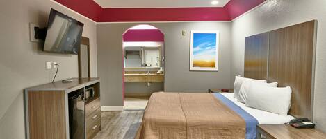 A hotel room with a bed, bedside table, desk, TV, and a bathroom with a mirror and sink.