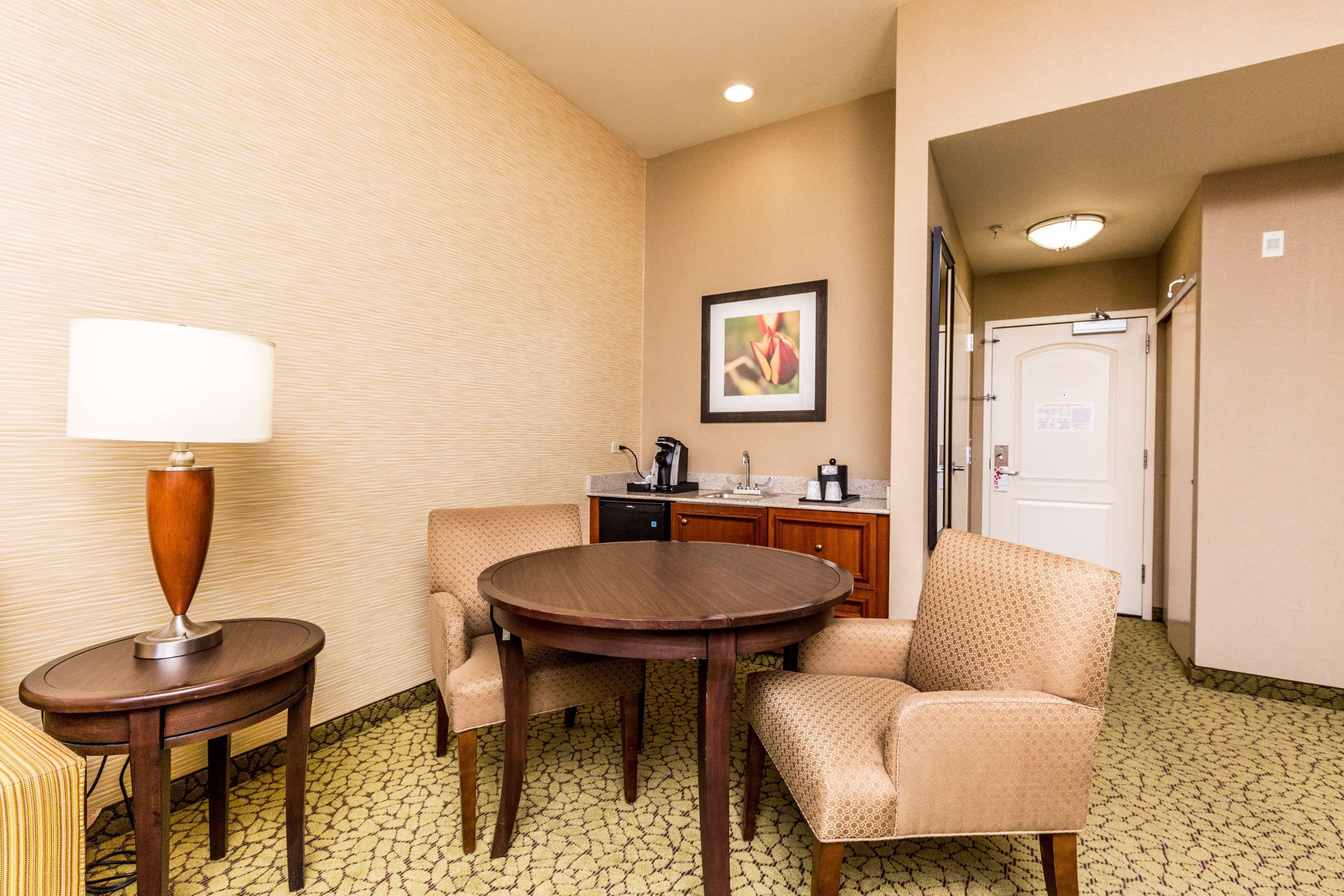 Habitación de hotel con una mesa redonda de comedor, dos sillones, una pequeña mesita con lámpara, una kitchenette y un cuadro en la pared.