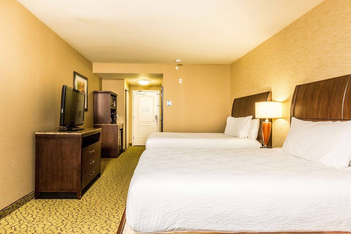 A hotel room with two beds, a TV on a wooden dresser, and a lamp on the nightstand.