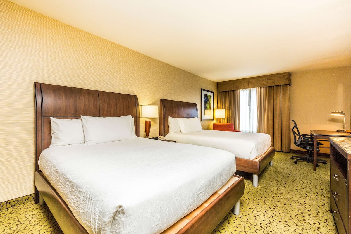A hotel room with two beds, a desk, a chair, and a lamp.