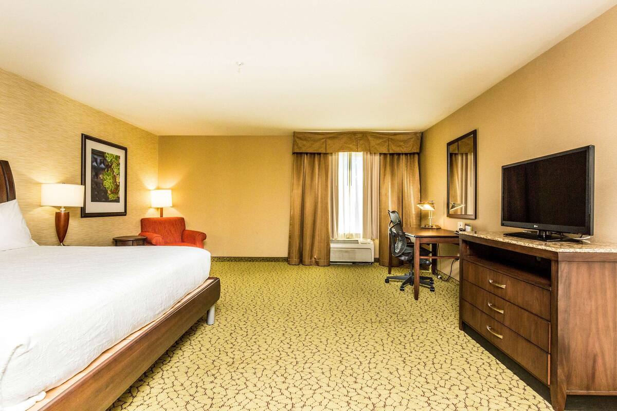 A hotel room with a bed, a desk with a television, a chair, and a desk lamp.