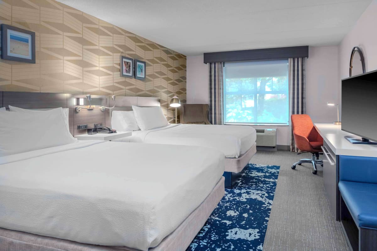 A hotel room with two beds, a desk, a chair, a TV, and a window with curtains.