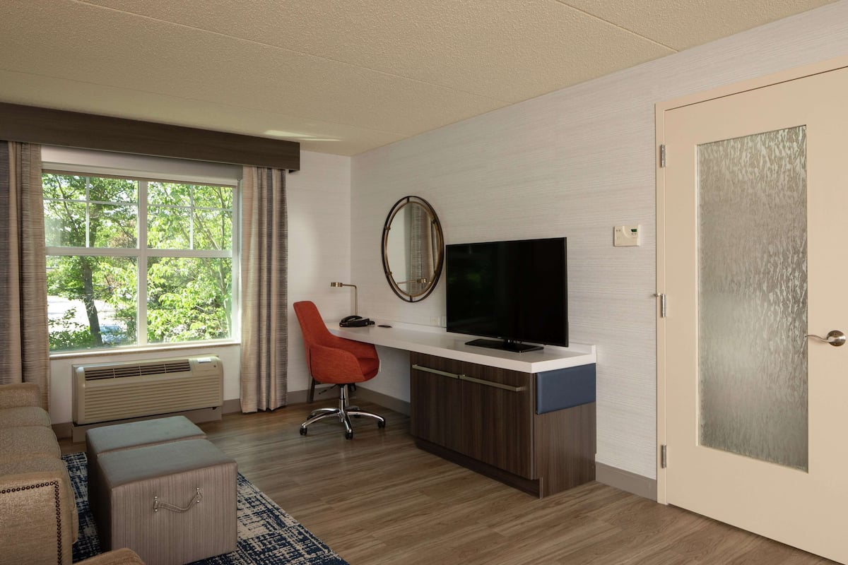 A modern hotel room with a large window, a desk with a flat-screen TV, a red office chair, and a wooden cabinet.