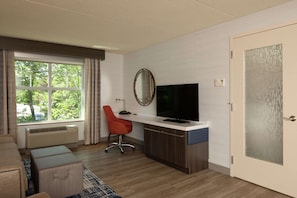 A modern hotel room with a large window, a desk with a flat-screen TV, a red office chair, and a wooden cabinet.
