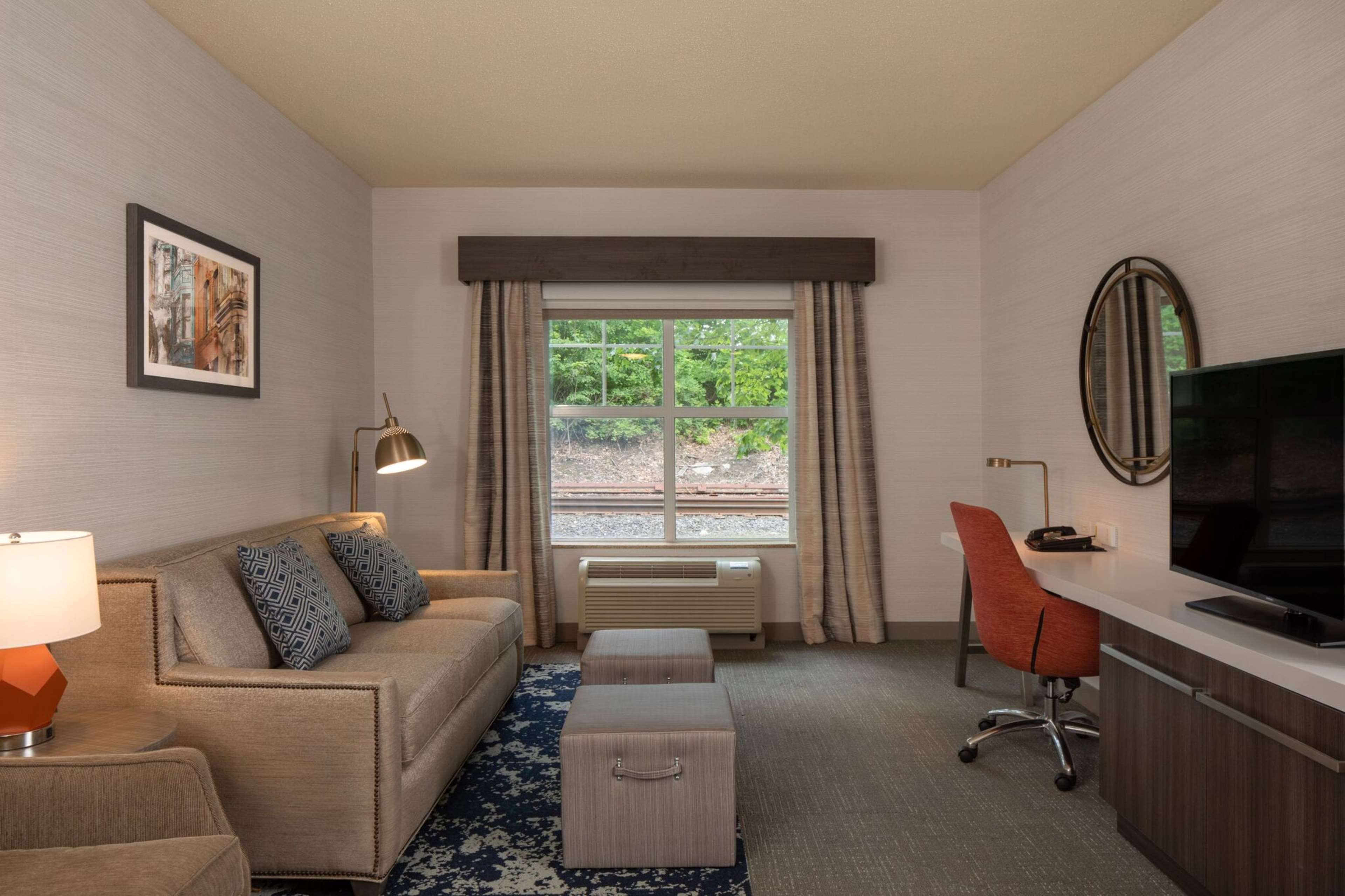 A hotel room with a sofa, ottoman, desk, TV, and a window with curtains.
