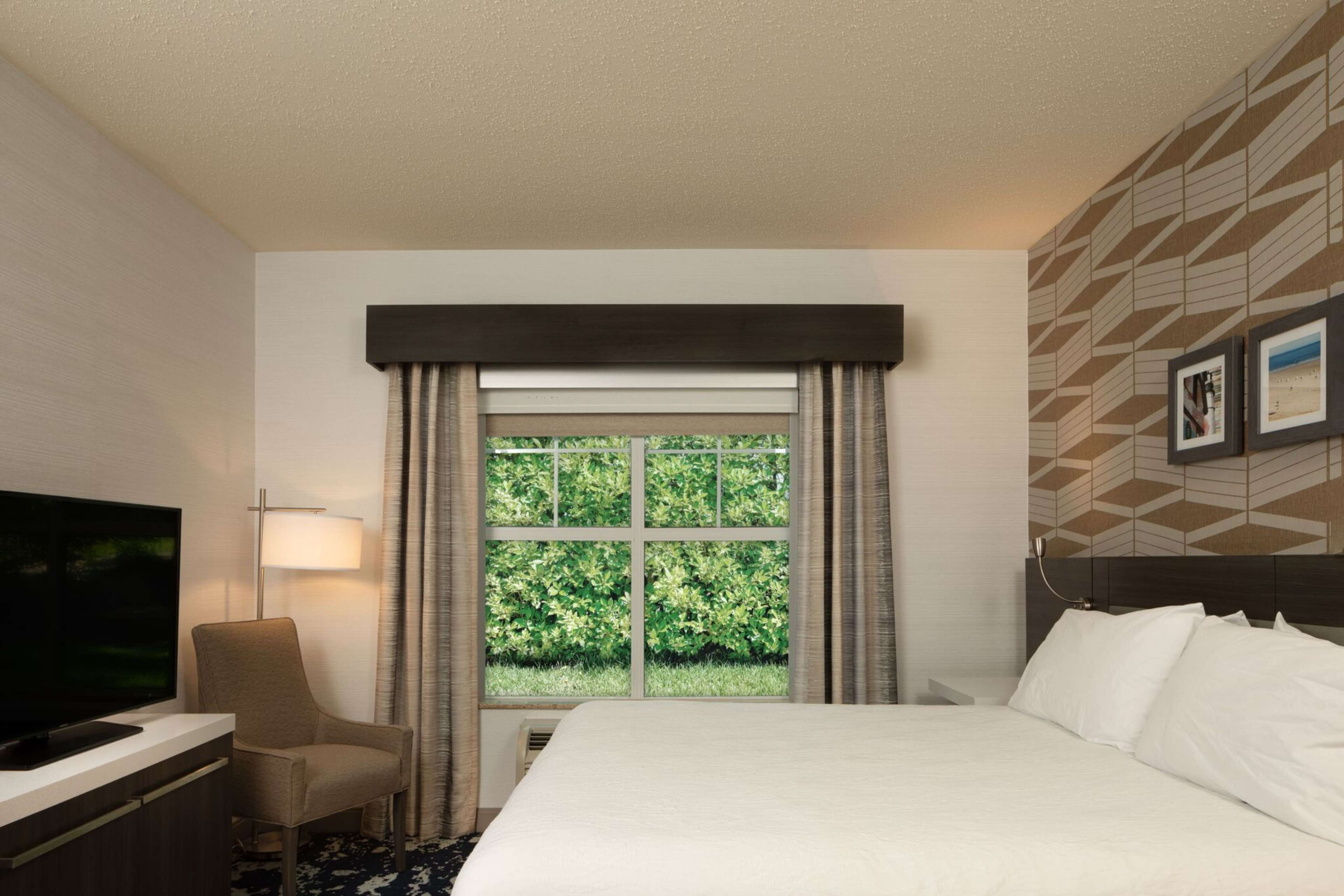 A hotel room with a bed, a TV, a chair, a window with curtains, and a wall with framed pictures.